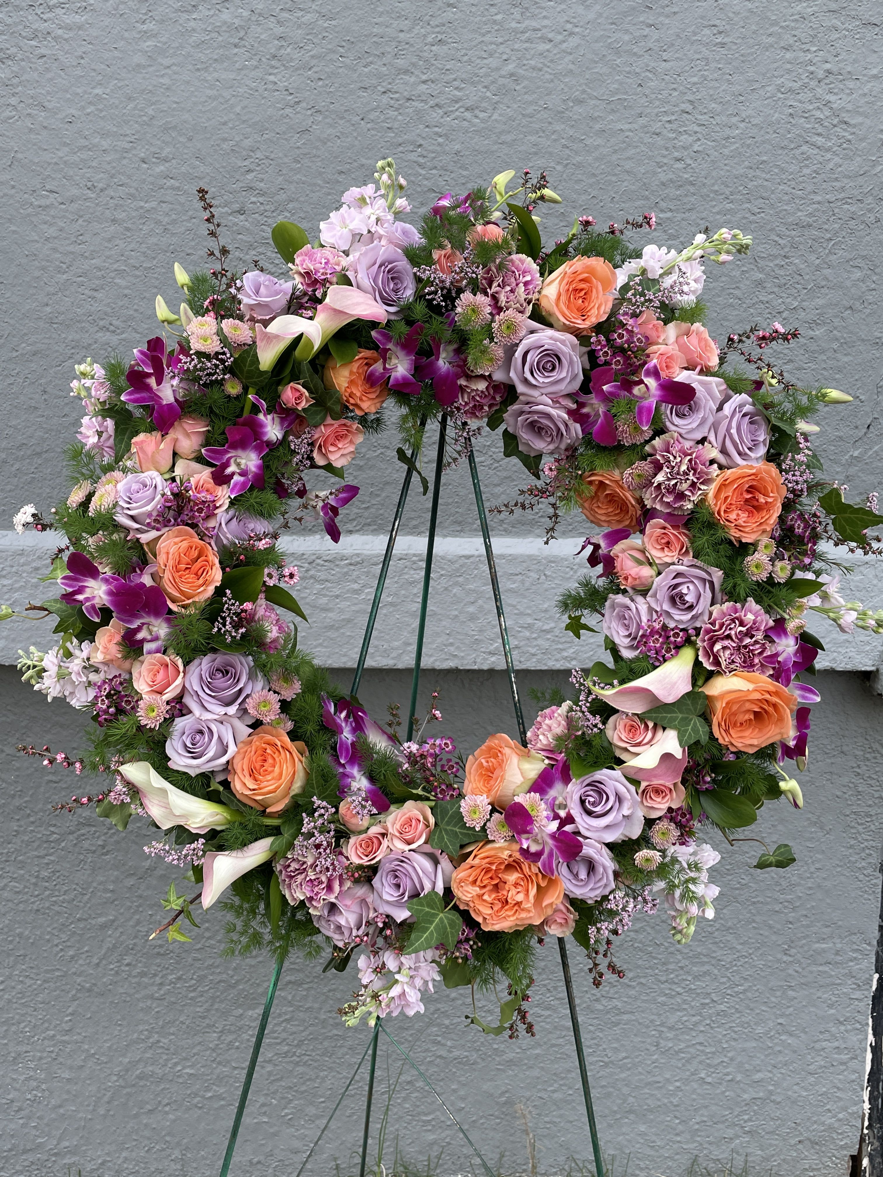 Custom Garden-Inspired Funeral Wreath Custom Garden-Inspired Funeral Wreath - MINIMUM 48 HOURS NOTICE FOR CUSTOM COLORS. A timeless expression of love, loss, and lasting honor, our Custom Garden-Inspired Funeral Wreath is a heartfelt tribute designed to celebrate a life beautifully lived. Measuring approximately 22 inches in diameter, this elegant floral wreath is handcrafted in a classic circular shape—symbolizing eternity and the unending cycle of life.  Lush, premium blooms and garden-inspired greenery are thoughtfully arranged to create a natural, flowing design that brings peace and gentle beauty to any memorial service, funeral ceremony, or graveside tribute. Whether displayed on an easel or placed at rest, this 22" wreath offers a blooming embrace during life’s most tender moments.  This design is fully customizable to reflect your personal vision. Please share your preferred color palette or floral inspiration, and our designers will create a one-of-a-kind arrangement tailored to your wishes. If no preferences are provided, we will select a seasonal and harmonious palette using our signature style.