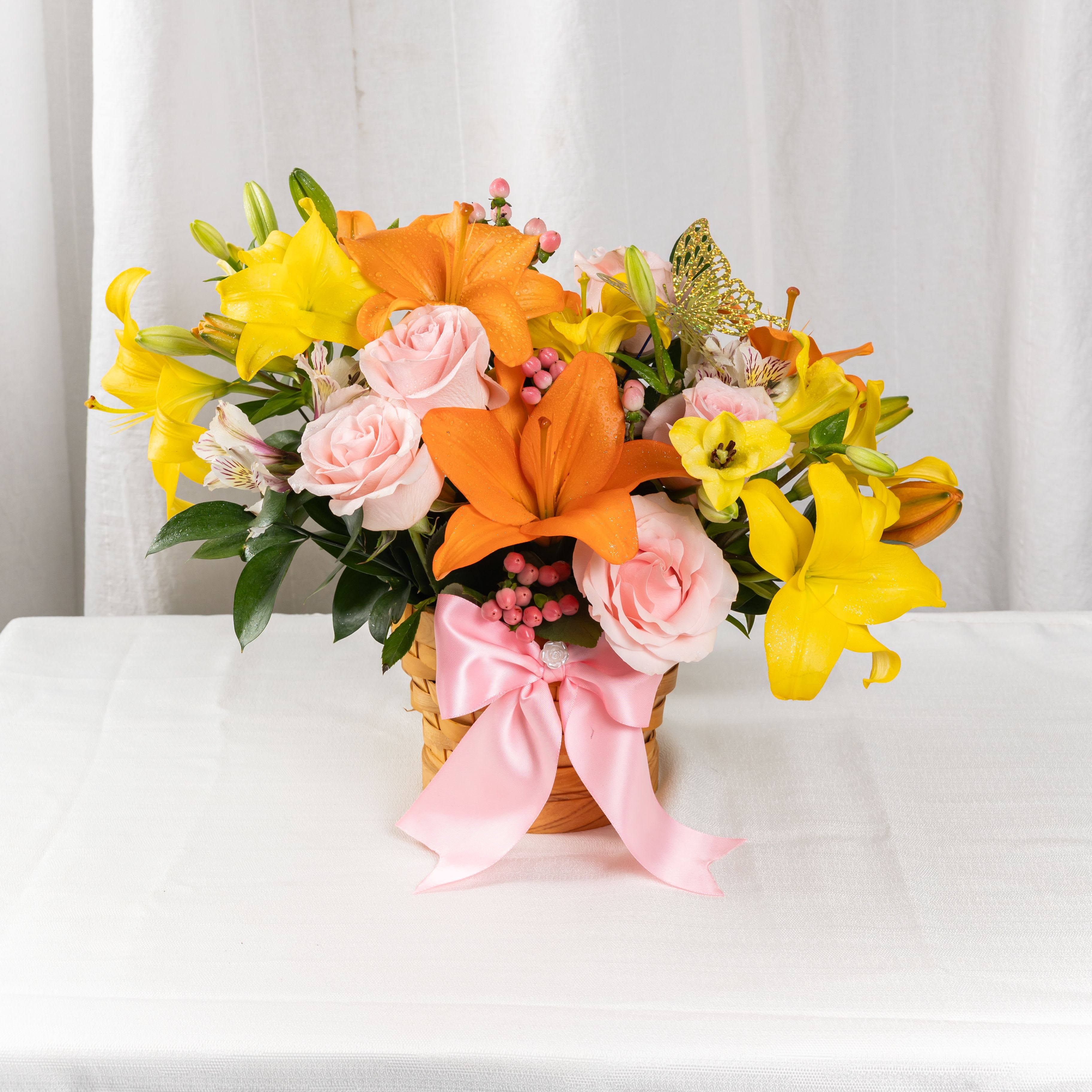 SPRING BREEZE - A cheerful basket of orange and yellow lilies, soft pink roses, and playful pink berries, tied with a satin ribbon and crowned with a butterfly. This radiant arrangement brings warmth, joy, and a touch of whimsy to any moment.