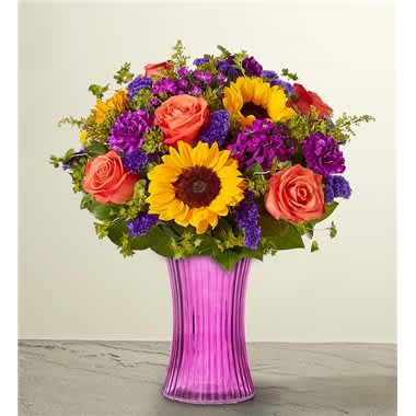 Floral Enchantment™ Bouquet - Set the scene for special moments with our striking arrangement. This enchanting floral mix is designed in our fluted violet gathering vase, bringing a bright surprise to celebrations of all kinds. • All-around arrangement with sunflowers, orange roses, purple dianthus, carnations and statice; accented with assorted greenery • Artistically designed in our exclusive fluted gathering vase in a lovely shade of violet; vintage pressed glass with ribbed detail creates a charming enhancement for the floral arrangement; measures 7.9&quot;H x 5&quot;D • Large arrangement measures approximately 17&quot;H x 14&quot;W • Medium arrangement measures approximately 15&quot;H x 13&quot;W • Small arrangement measures approximately 14&quot;H x 12&quot;W • Our florists hand-design each arrangement, so colors and varieties may vary due to local availability • Sunflowers may arrive with light or dark centers