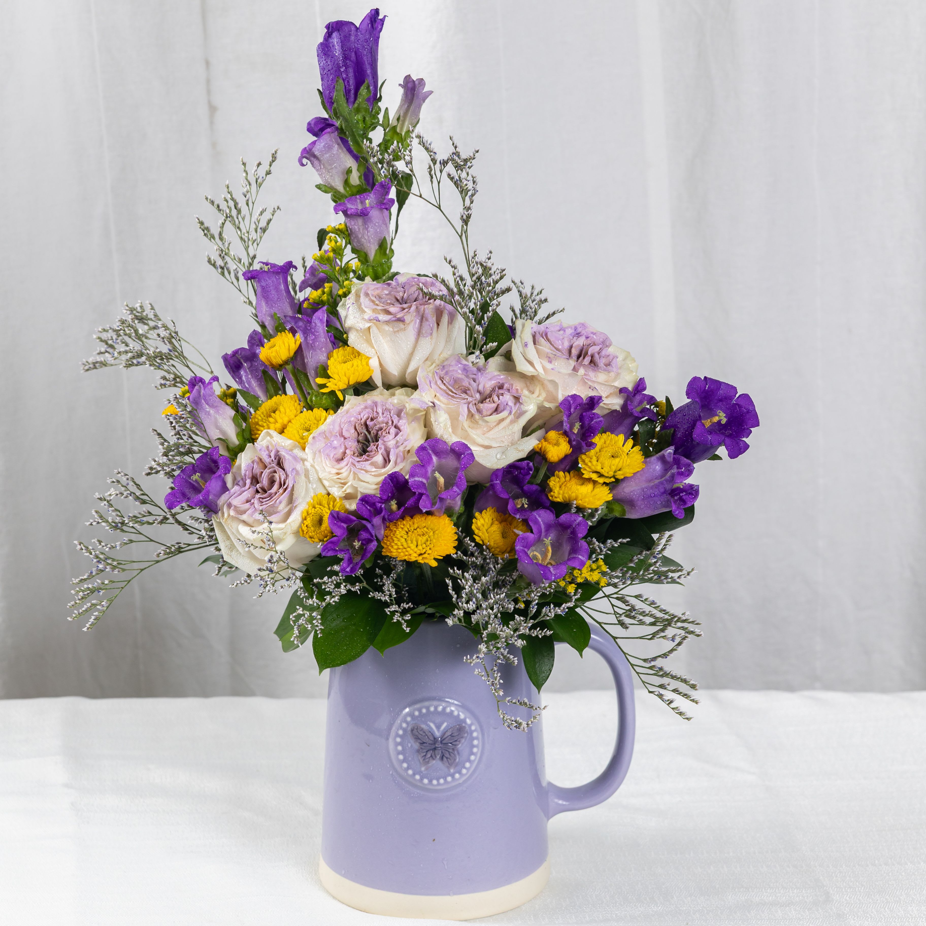 Bright Day - A dreamy mix of soft purple roses, bell-shaped blooms, yellow button flowers, and airy baby's breath, arranged in a ceramic pitcher adorned with a butterfly. This delicate bouquet evokes charm, serenity, and a touch of countryside magic.