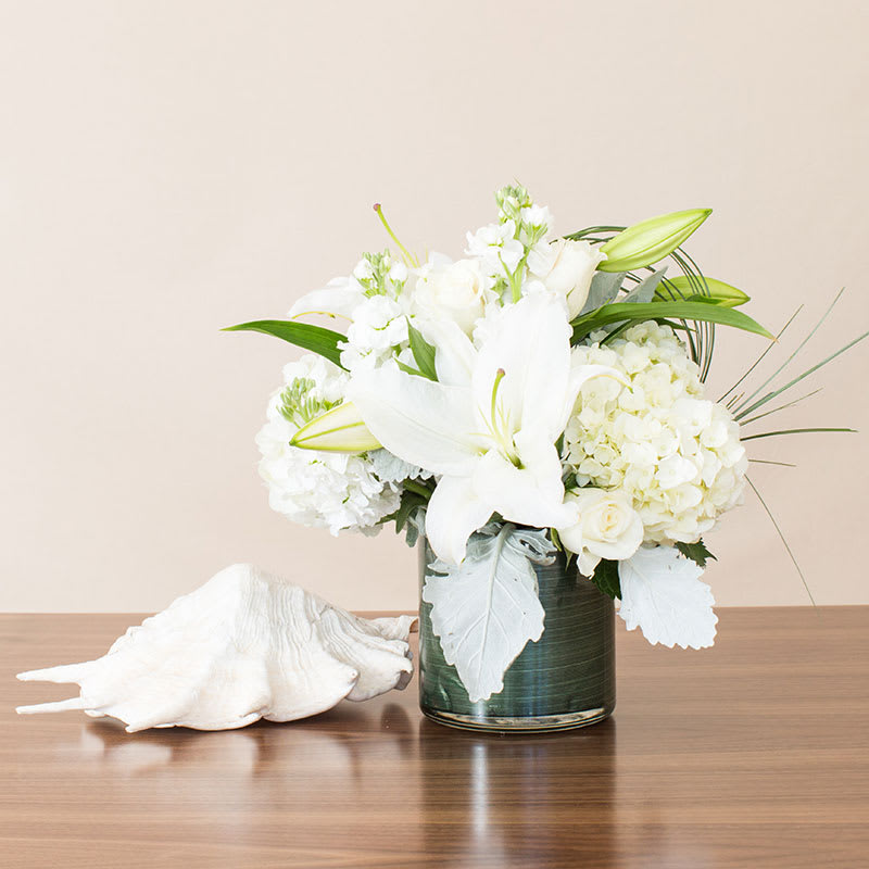 Pure Elegance - Capture the spirit of graceful gardens with our mixed arrangement of popular white garden flowers. This elegant display features hydrangeas, roses, and lilies presented in a glass vase, creating a serene and enchanting ambiance.  Key Features:  White Garden Flowers: Includes a mix of popular white garden flowers such as hydrangeas, roses, and lilies, symbolizing purity and elegance.  Glass Vase: Presented in a sleek glass vase, enhancing the beauty and visibility of the flowers.  Graceful and Elegant: Evokes the spirit of graceful gardens, combining freshness and natural beauty in every bloom.  Perfect for Any Occasion: Ideal for birthdays, anniversaries, or as a heartfelt gesture to express sympathy or appreciation.  Serene Ambiance: Creates a serene and enchanting ambiance, perfect for enhancing any space with its timeless charm.  Celebrate the beauty of nature with our Mixed White Garden Flowers in Glass Vase, a perfect choice for conveying tranquility and elegance with its exquisite blend of garden blooms.