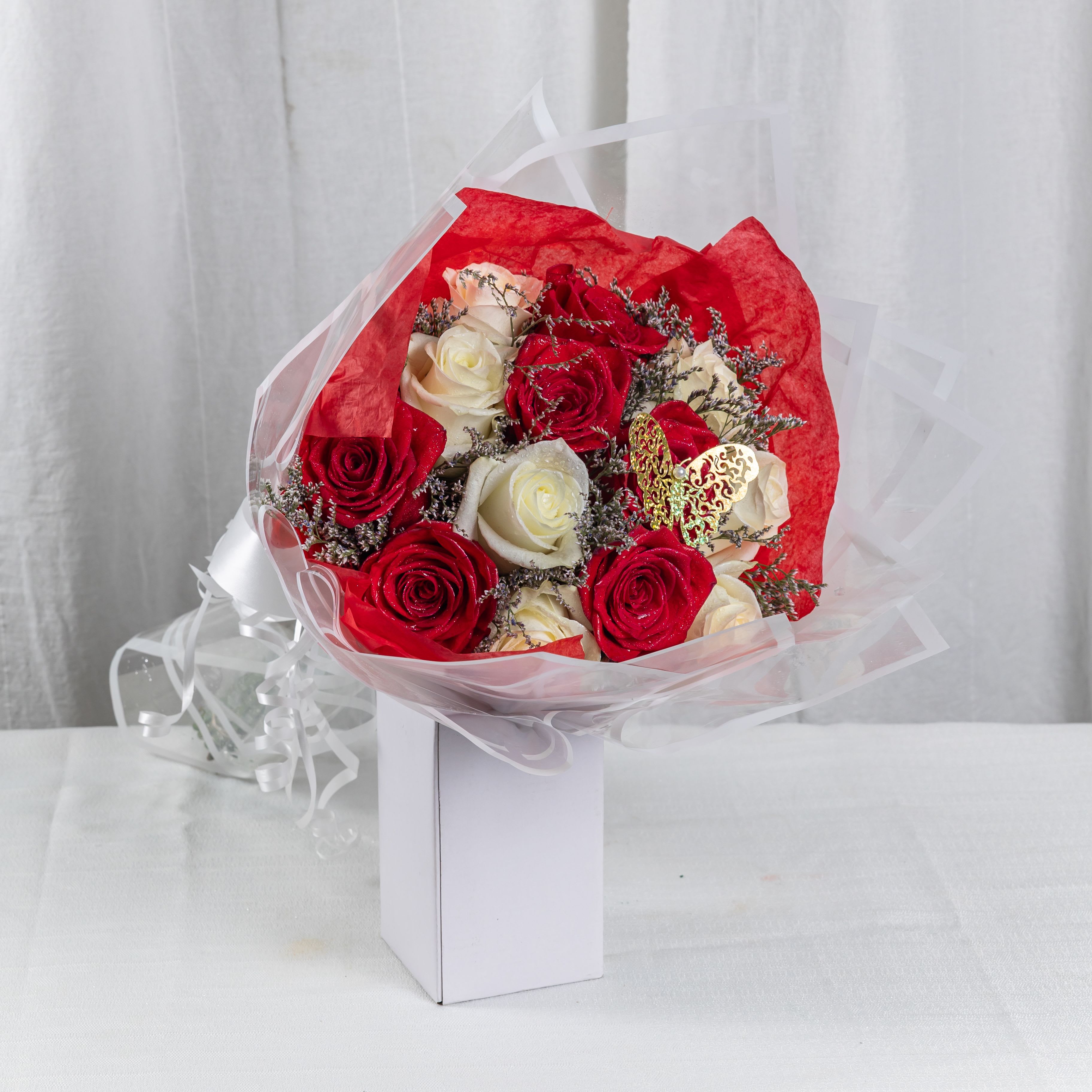 Butterfly Embrace (12 Roses) - A romantic blend of red and white roses, accented with delicate purple filler blooms and wrapped in vibrant red and sheer cellophane. Nestled in a white box and adorned with a golden butterfly and pearl-tied ribbon, this bouquet is a graceful celebration of love, elegance, and whimsy.