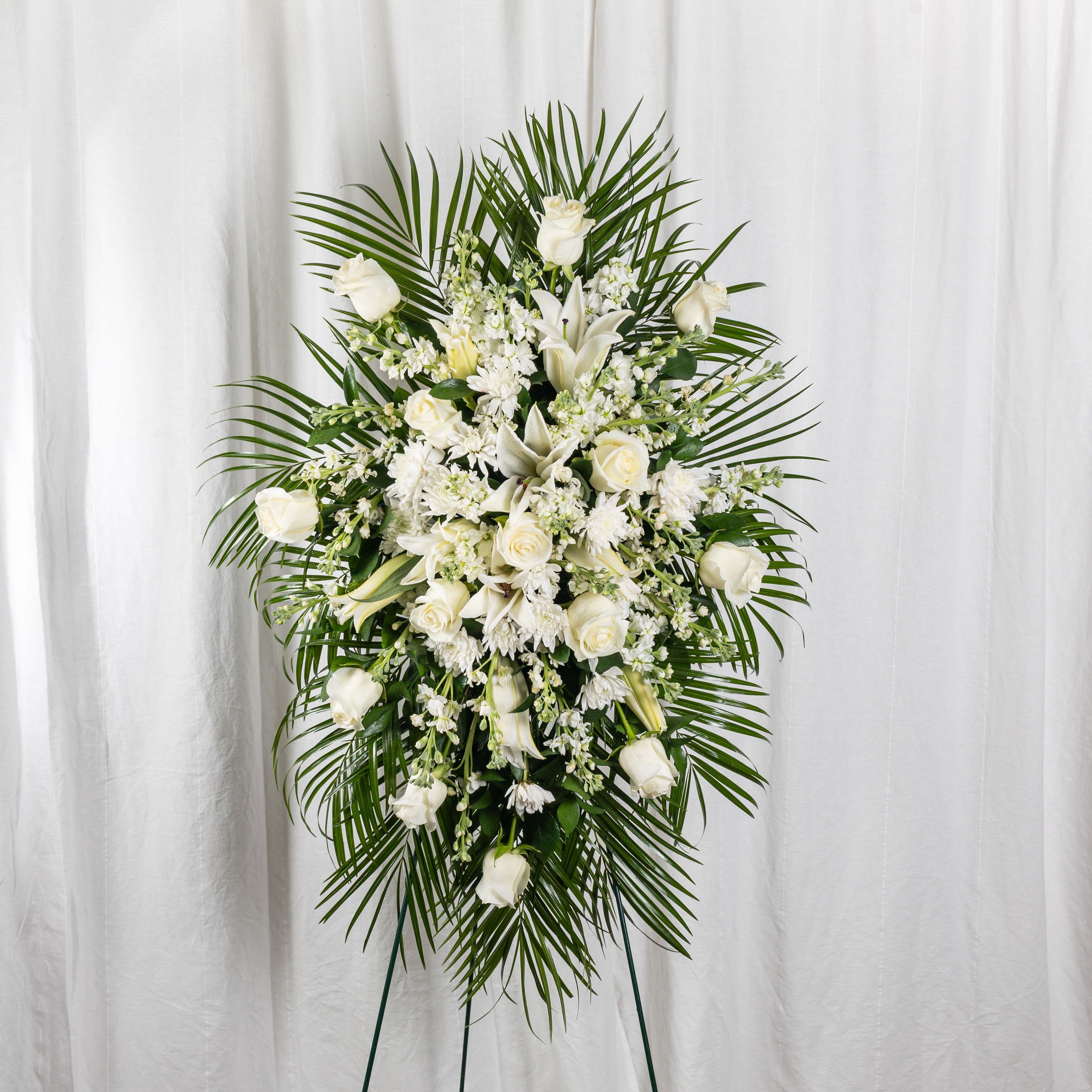 REST IN PEACE - An elegant standing spray of white roses, lilies, and delicate blooms, framed by lush palm leaves and set against a soft white backdrop. This graceful tribute speaks of peace, purity, and heartfelt remembrance—honoring life with reverence and beauty.