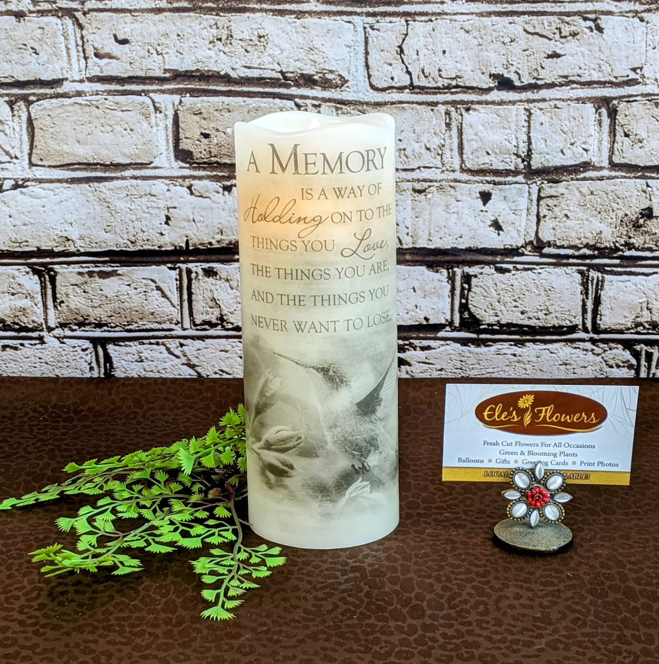 Memory - Sympathy Flicker Candle Memory - Sympathy Flicker Candle - Offer a gentle and enduring symbol of comfort with our Flameless Flicker Sympathy Pillar Candle. This elegant candle features a realistic, flickering LED flame, providing a warm and soothing glow without the worry of an open flame. Encased in a classic pillar design, it serves as a beautiful and safe tribute during times of remembrance and sympathy. Ideal for memorial services or as a thoughtful gift, this flameless candle is a lasting expression of support and solace.  Candle comes printed with the phrase, "A memory is a way of holding on to the things you love, the things you are, and the things you never want to lose." Uses 2 AA batteries, included.   We are able to incorporate this into a floral display or plant. Please inquire with us.   Approximate Dimensions: 8"H x 3"W
