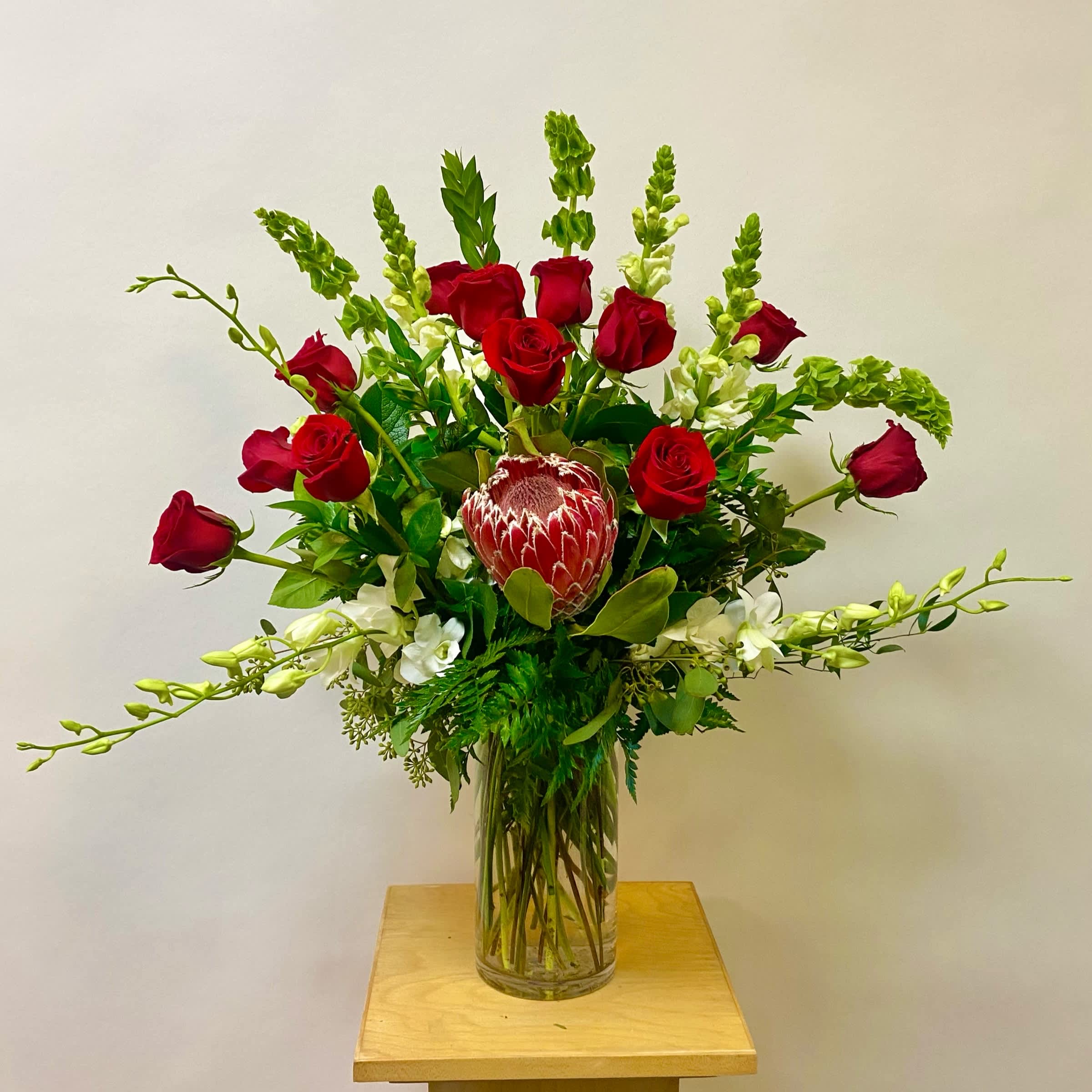 Eye Candy Eye Candy - This Tall Arrangement includes a Dozen Red Roses, White Snapdragons, Bells of Ireland, White Dendrobium Orchids, Wax Flower and Pink Protea with Greenery in a Glass Vase. Or as Similar as Possible Depending on Stock Availability.