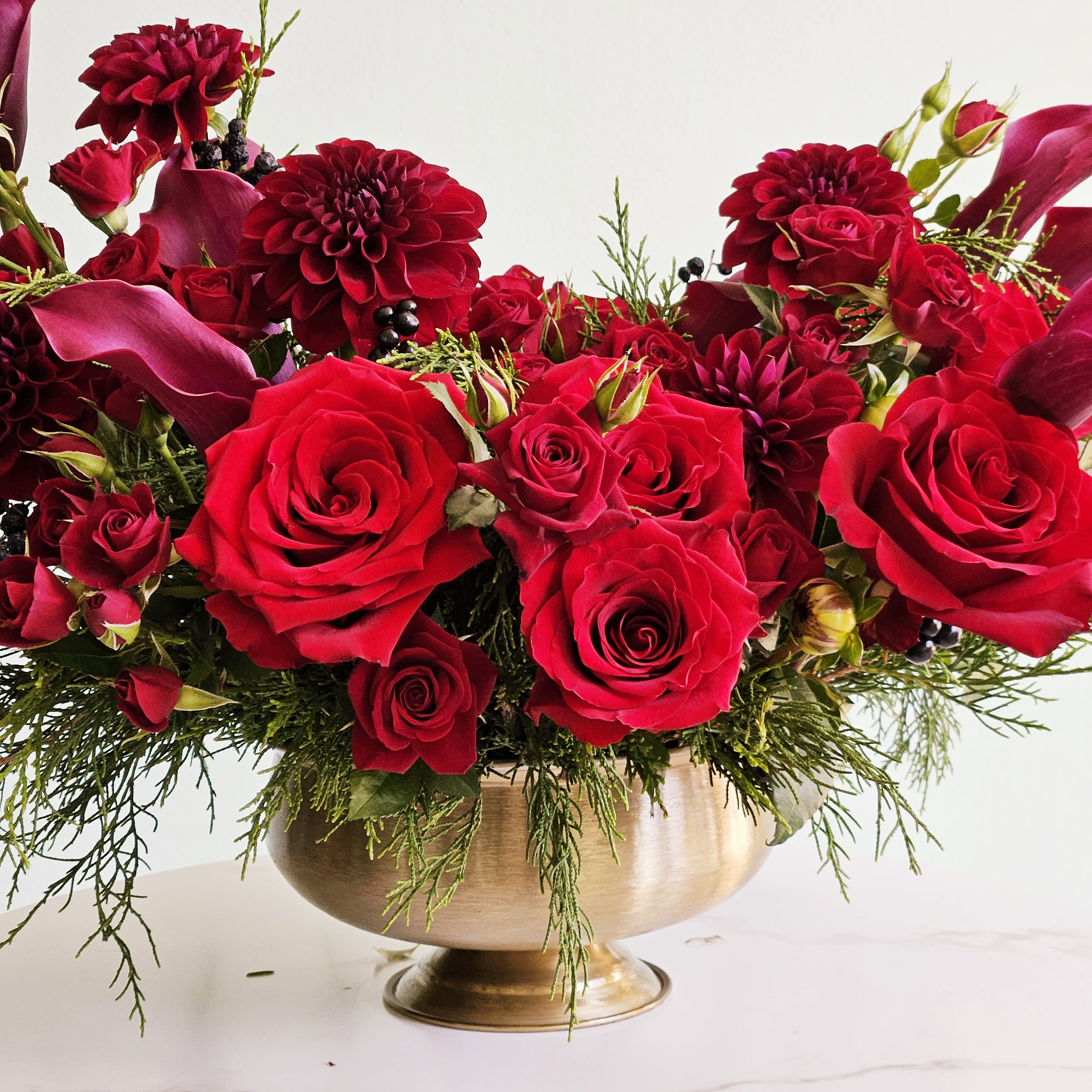 Golden Merlot  - Indulge in the opulence of our Golden Merlot arrangement. Featuring rich burgundy roses, dahlias and calla lilies nestled in a gleaming gold vase, this stunning design exudes romance and refinement. Perfect for marking life's most memorable moments or adding a dramatic flair to any space, its deep hues create an unforgettable statement. Gift this luxurious arrangement to express heartfelt sentiments or adorn your home with its timeless beauty.  Please note: The arrangement pictured is our deluxe version, showcasing extra fullness and blooms for a more luxurious presentation.