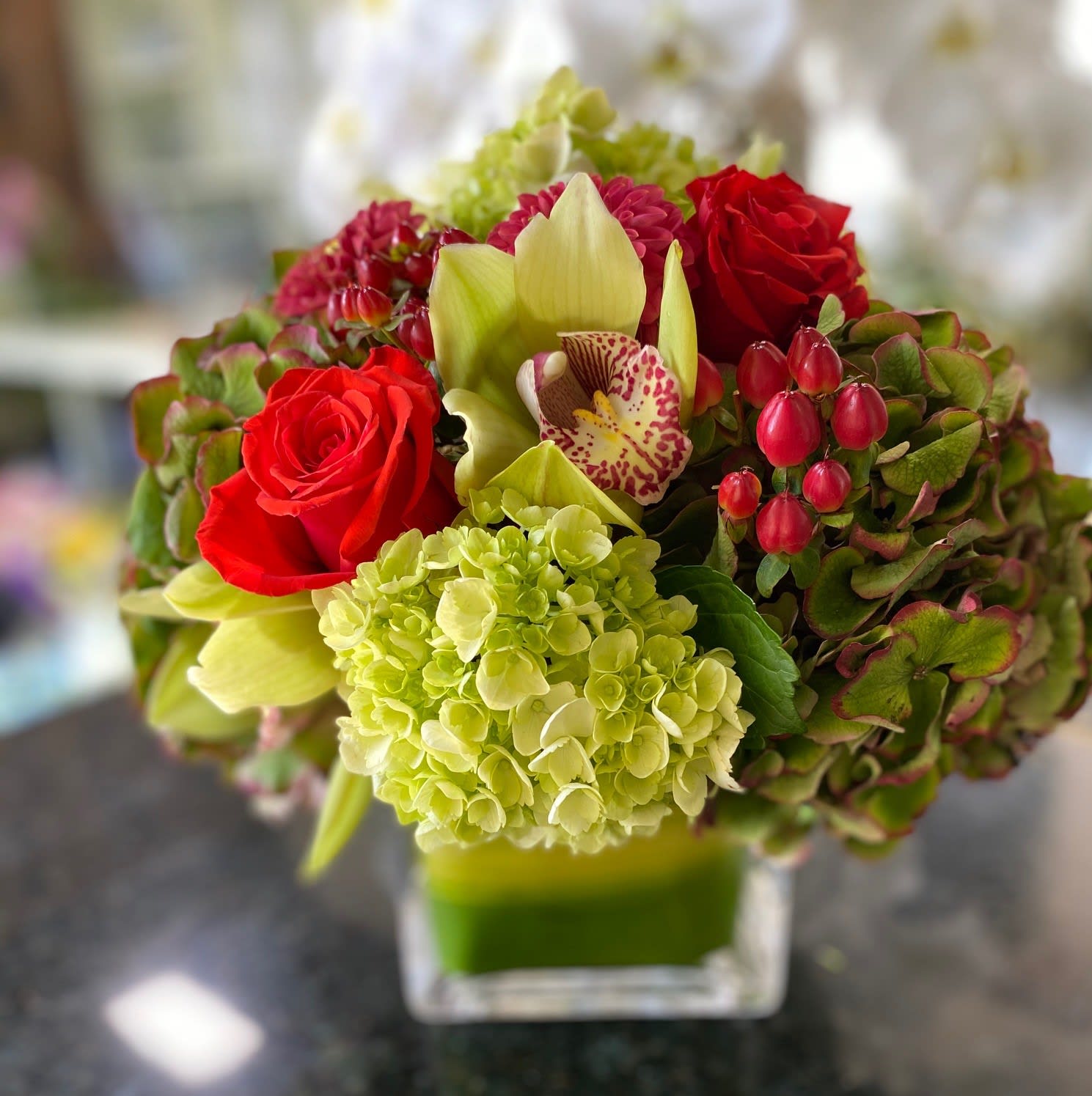 A Touch of Fall - The perfect table centerpiece ........Perfect for that table setting. With Beautiful Fall colors , including Dahlias ,Dutch Hydrangea, Hypericum with some Cymbiduim Orchids for that oh so special look. Standing about 9-10 inches tall and made in a 5X5 glass Cube carefully lined with a leaf.