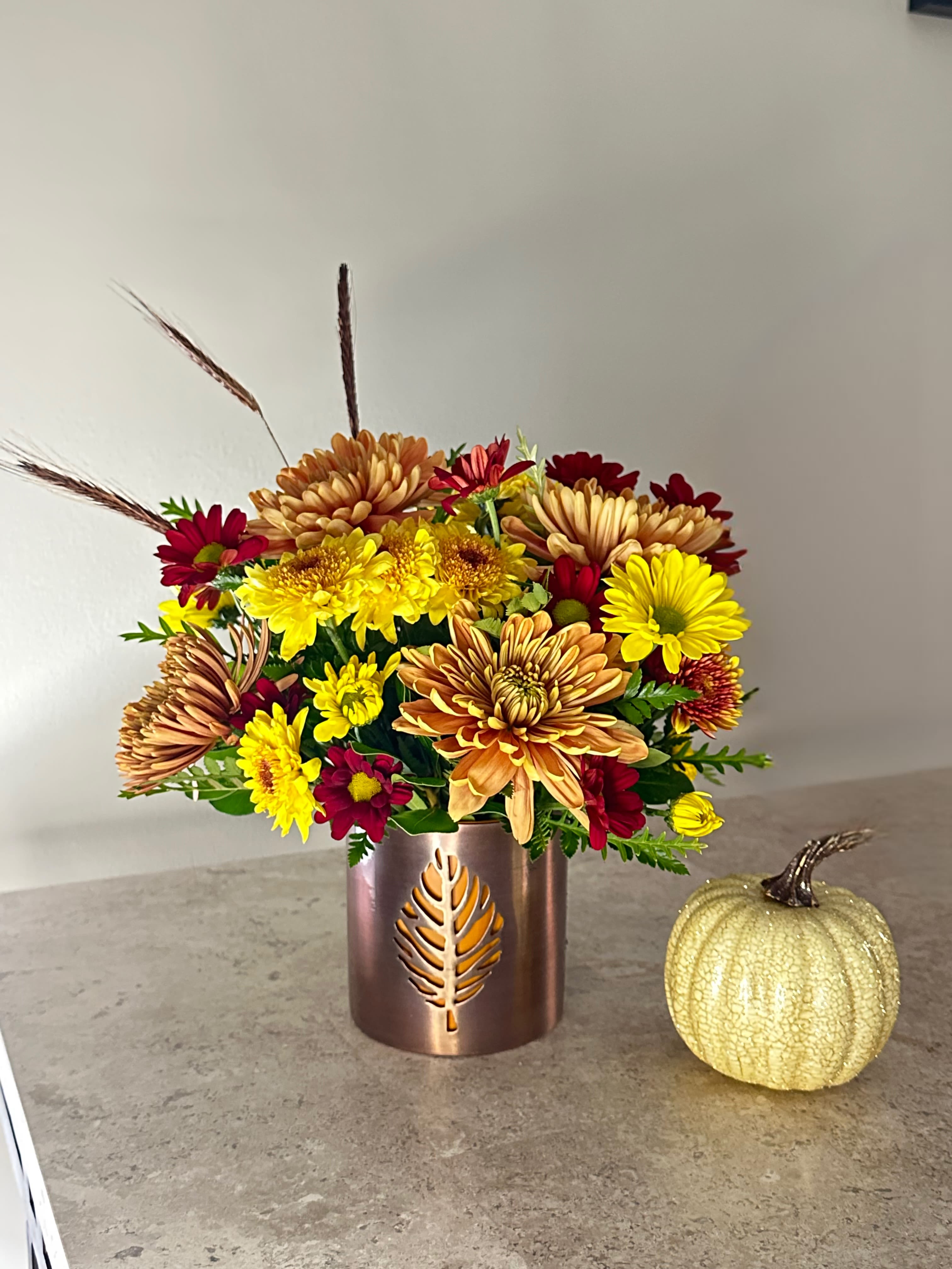 Autumn Glow Bouquet Autumn Glow Bouquet - Bring the warmth of fall indoors with our Autumn Glow Bouquet. Bursting with vibrant yellow, burnt orange, and deep burgundy mums, this seasonal arrangement is accented with wheat stems for a rustic harvest touch. Designed in a copper leaf-cut vase that glows beautifully with candlelight, it’s the perfect accent for your home, Thanksgiving table, or as a thoughtful autumn gift. Paired with its cozy charm, this bouquet celebrates the beauty of the season in every bloom. small pumpkin not included.