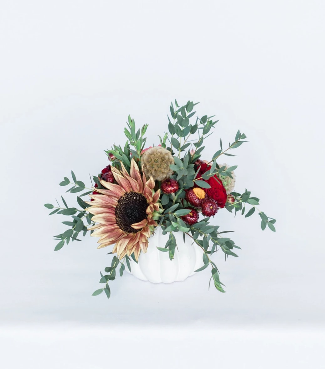 Rustling Roses - Celebrate the changing of the seasons with our new Fall Collection! Inspired by the rustic warmth that autumn brings, the Rustling Roses comes arranged with a beautiful mix of deep red and orange premium blooms and textures, all eloquently designed in one of our hand-picked designer pumpkin vases.**  Pictured [STANDARD]  *Some items in this arrangement must be special ordered, please allow 1-2 weeks notice to allow time for arrival of special items, otherwise, substitutions will occur.*  **While supplies last. Vase will be substituted with another designer vase once the pumpkin vase is out of stock. Please contact the studio directly for any questions.