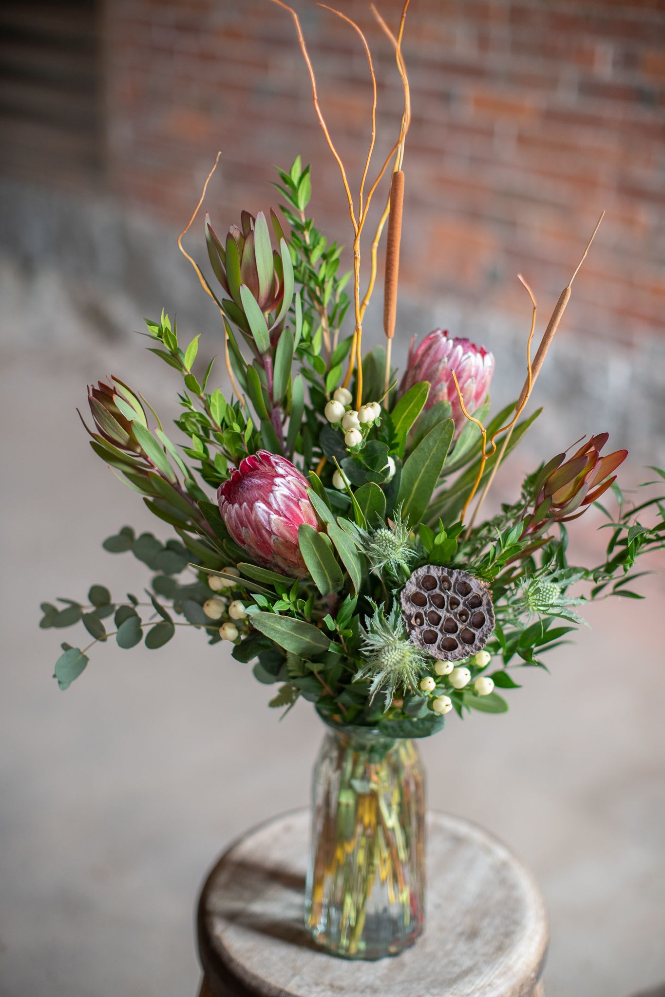 Princess Vase - Nothing says &quot;Pretty, pretty princess,&quot; more than unique tropical protea flowers. These exotic blooms are native in Australia, and South Africa. They presently are commercially grown in Hawaii and California also. These rare flowers are perfection in soft pink to mauve hues. They're paired with safari sunset Leucadendron in a rich red tone. Autumn pencil cat tails, preserved lotus pods, thistle, and hypericum berries and textural elements that elevate this design. All artfully arranged with seasonal foliage in a tall cinch style vase. Original deign by Kate - Orientation is one sided. In some instances we may need 24-48 hour lead time to order exotics - we do our best to keep out of stock items accurately marked. 