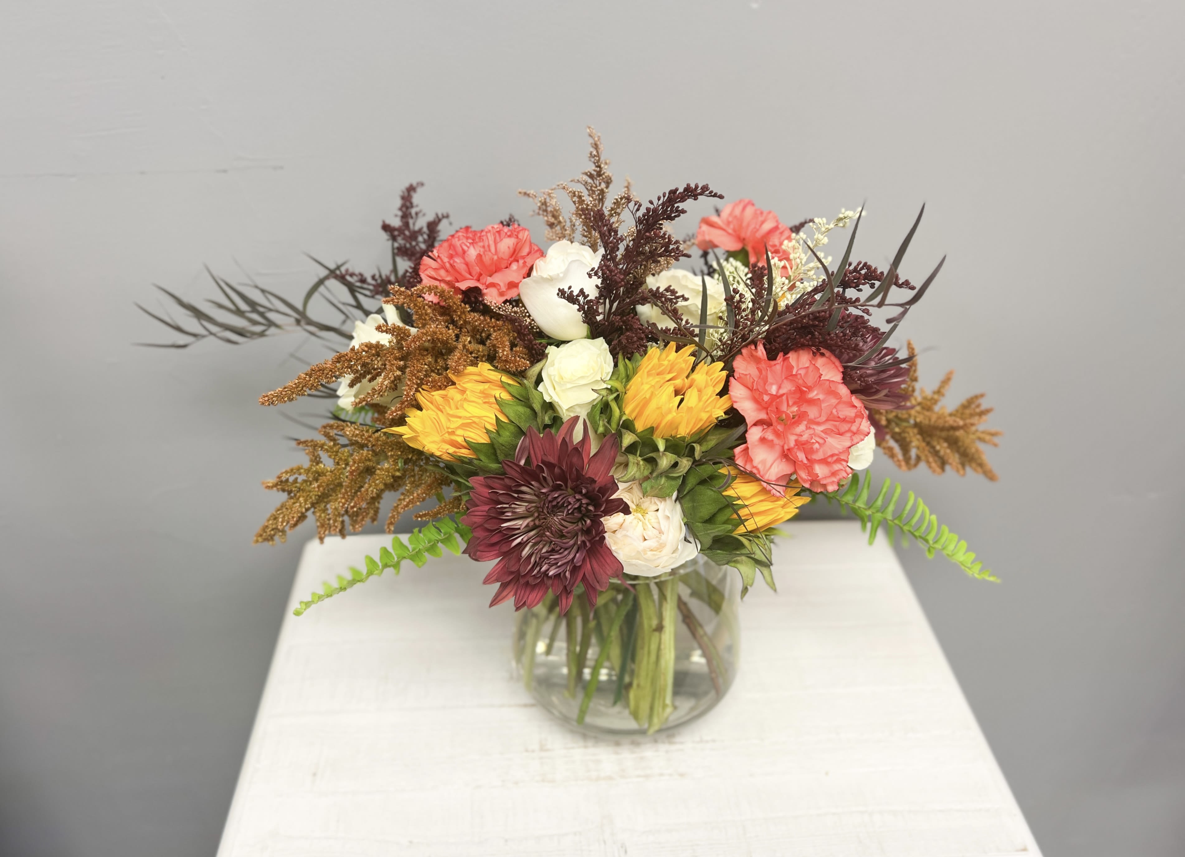 Golden Sunset  - Bring warmth and charm into any space with this vibrant fall-inspired arrangement. Featuring bold sunflowers, elegant roses, coral carnations, and textured foliage, this design radiates seasonal beauty. Arranged in a clear glass vase, it’s the perfect gift for birthdays, celebrations, or simply brightening someone’s day.  Each bouquet is hand-designed with fresh blooms, so variations in flower types and colors may occur, making every arrangement uniquely beautiful. 