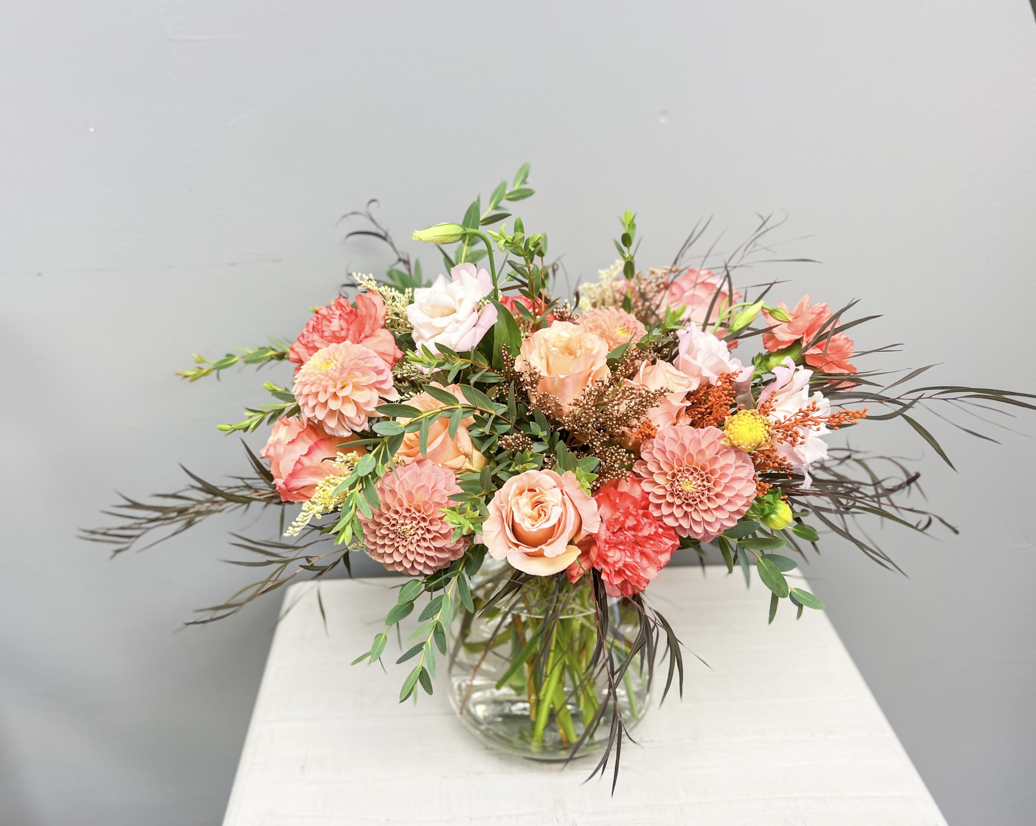 Coral Charm - Soft romance meets timeless elegance in this lush garden-style arrangement. Featuring peach and blush roses, coral carnations, delicate lisianthus, and perfectly rounded dahlias, accented with seasonal textures and greenery, this bouquet radiates warmth and charm. Designed in a clear glass vase, it makes a stunning gift for anniversaries, birthdays, or simply sending love.  Each arrangement is hand-crafted with fresh seasonal blooms, ensuring every piece is unique and one-of-a-kind.