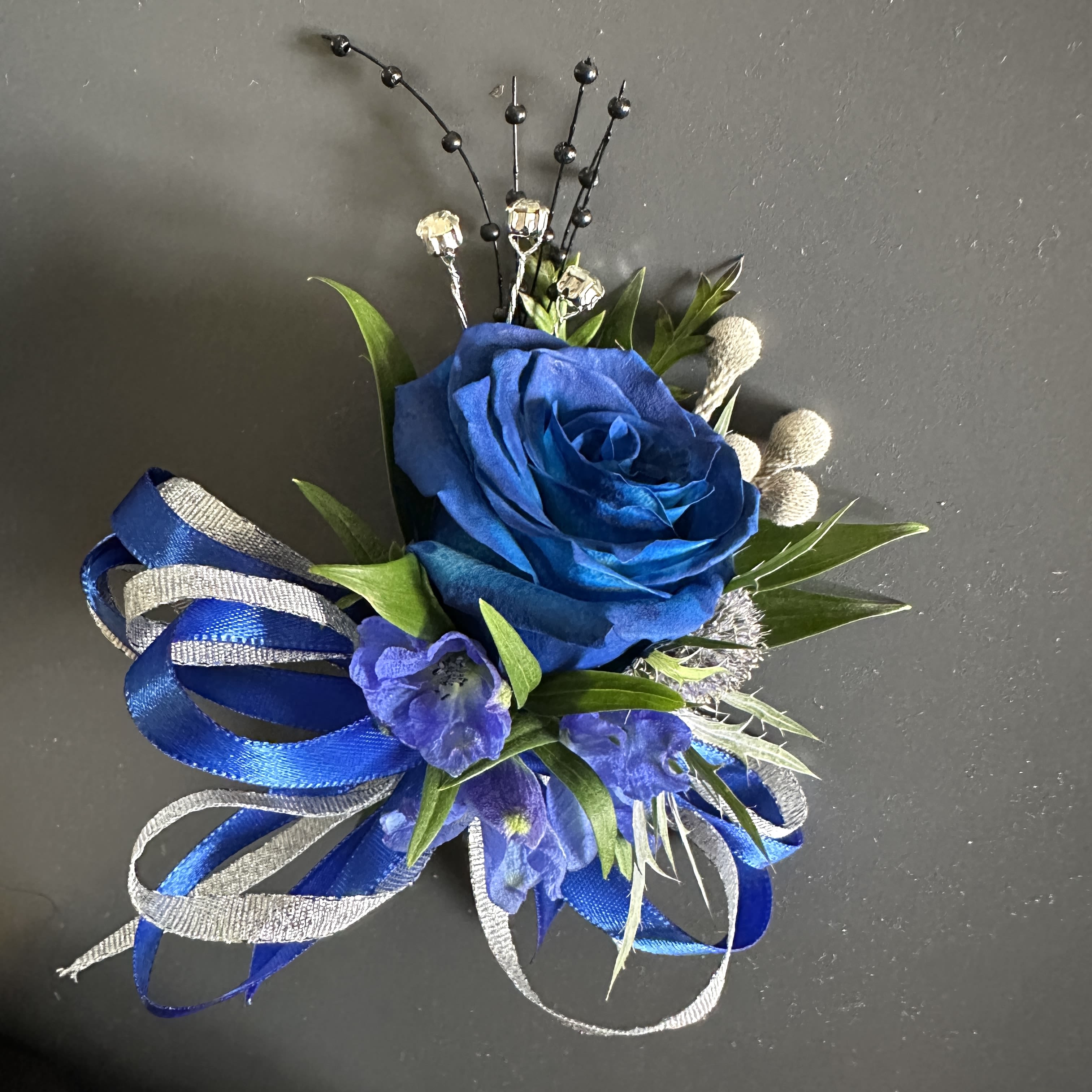 Royal Blue Corsage - Royal Blue Elegance Corsage This striking pin-on corsage features a vibrant blue rose accented with delicate purple blooms, lush greenery, and silver details. Finished with rich blue and shimmering silver ribbons, it brings a touch of sophistication and luxury—perfect for weddings, formal events, or special celebrations. 