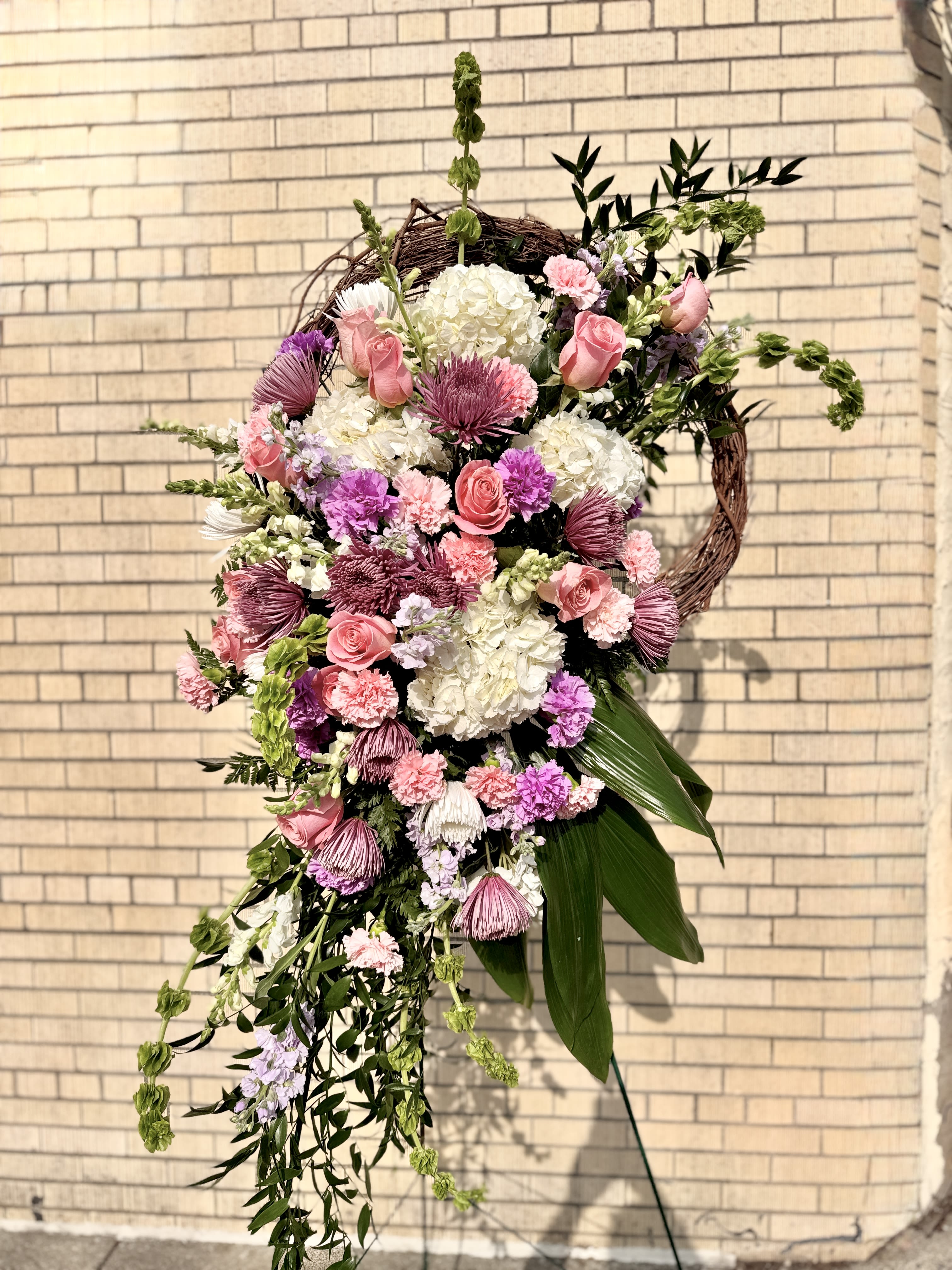 Lavender Compassion Standing Spray - Delicately adorned with an array of Lavender and  pink blooms, including roses, carnations, and snapdragons, this standing spray is a steady display of love and devotion. Each petal whispers of empathy and understanding, offering comfort and support in moments of sorrow. Order Lavender Compassion today and let us create something to honor your loved one. 