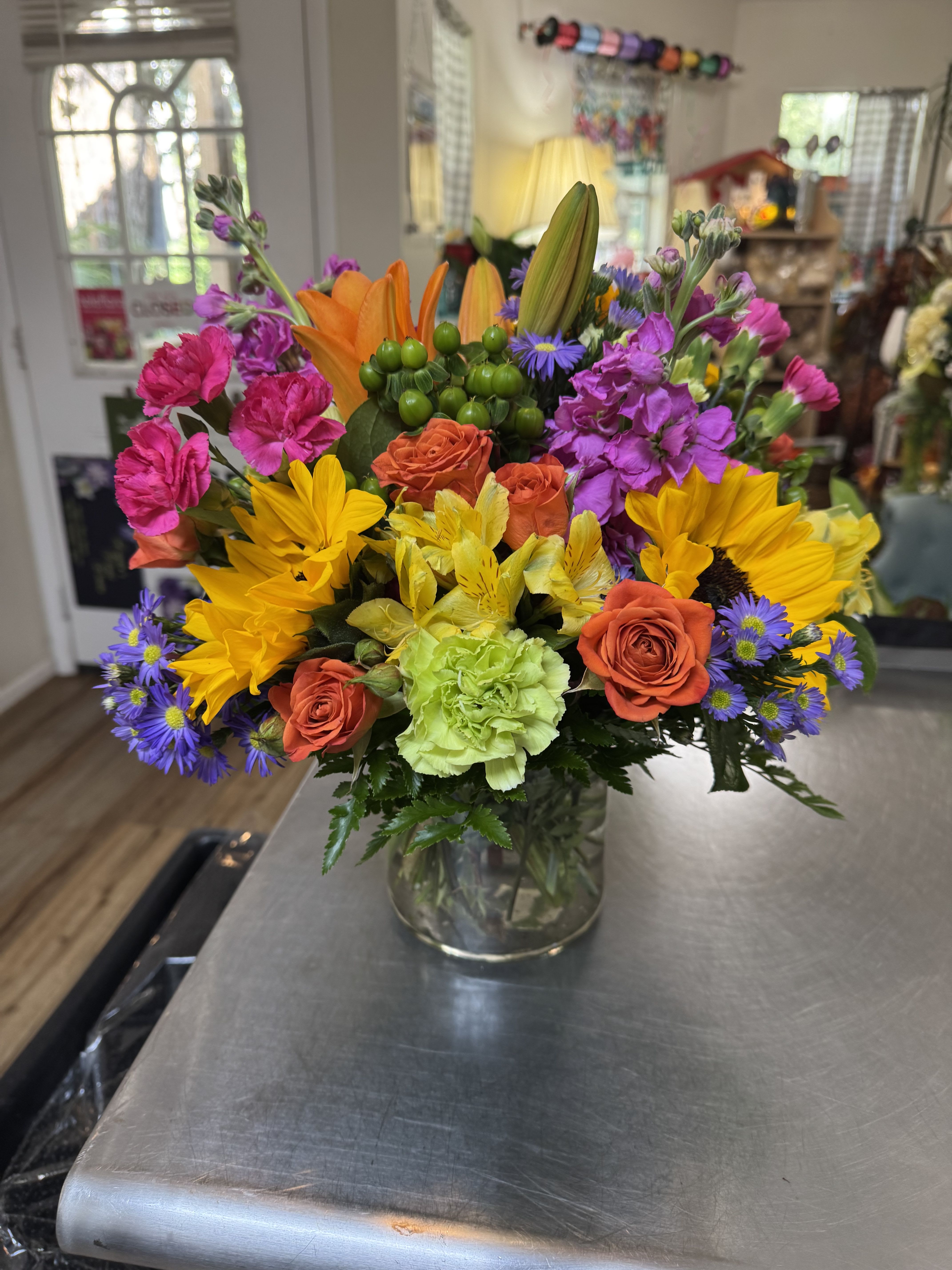 Sunburst Garden Bouquet - Celebrate the joy of color with our Sunburst Garden Bouquet, a cheerful and vibrant floral arrangement designed to bring warmth and happiness to any occasion. Bursting with bold orange roses, sunshine-yellow daisies, bright pink carnations, green carnations, purple asters, and elegant lilies, this bouquet is a stunning showstopper in a clear glass vase.  Perfect for birthdays, get well wishes, congratulations, or just because, this handcrafted arrangement is available for same-day flower delivery in Pierson and Deland, FL.   Designed by expert florists at Pierson Florist and Deland Florist  Bright, bold, and full of life – ideal for celebrations or everyday joy  Fresh, hand-picked blooms arranged in a stylish glass vase  Fast, reliable flower delivery in Pierson and Deland – satisfaction guaranteed  Bring a splash of color into someone’s life today with trusted local Pierson and Deland flower delivery service. Order now and let your sentiment shine through flowers!