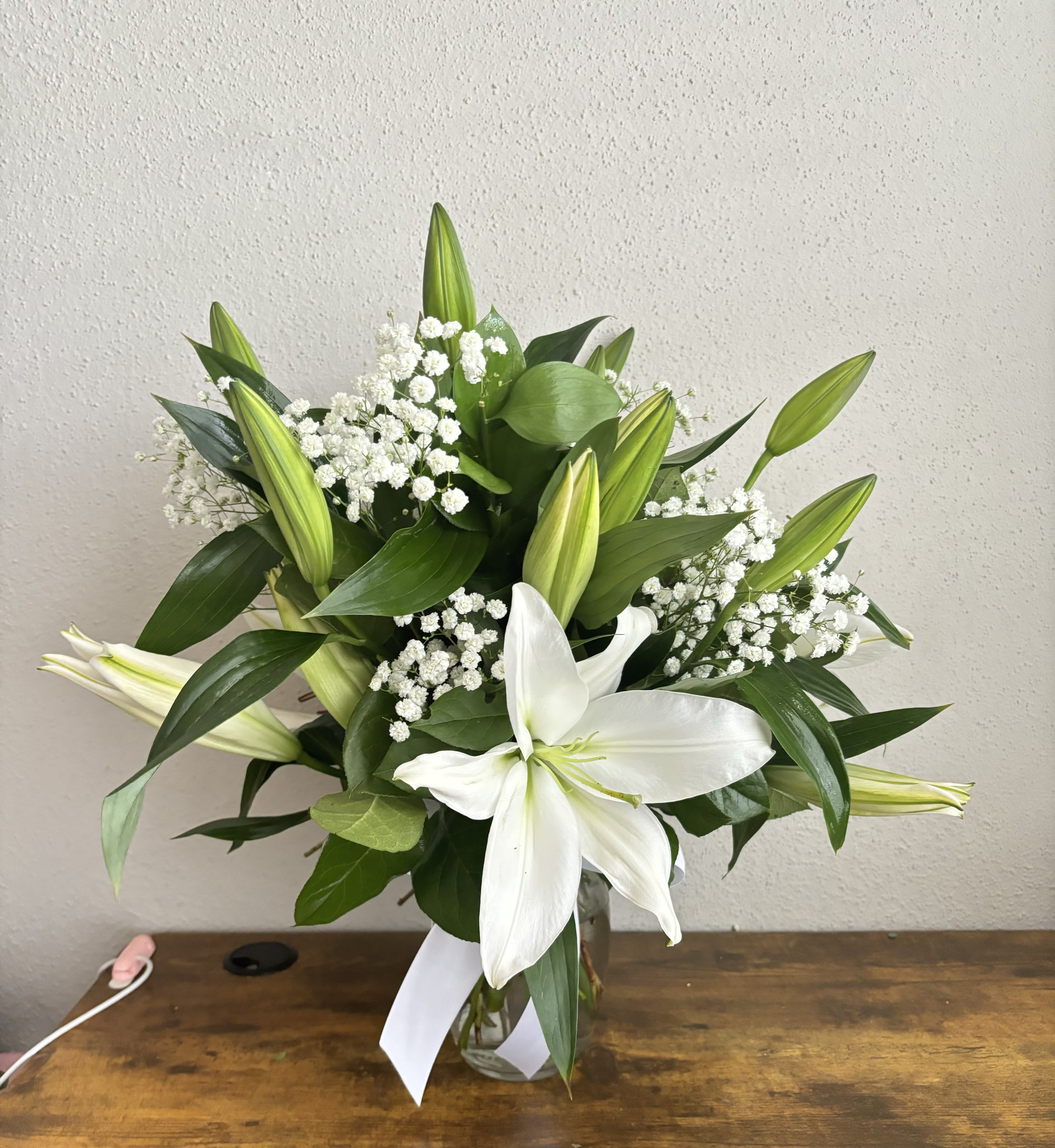 White Lily Elegance Vase Arrangement - White Lily Elegance Vase Arrangement  Bring timeless beauty and calm sophistication to any space with our White Lily Elegance Vase Arrangement. This graceful bouquet features pristine white lilies, artfully accented with lush aspidistra leaves and elegant lily foliage for a bold yet balanced look. Arranged in a sleek glass vase, it’s a refined statement of purity and serenity.  Perfect for expressing sympathy, celebrating milestones, or simply brightening a room, this arrangement radiates peace, grace, and natural charm. Its soft fragrance and minimalist elegance make it ideal for both home décor and heartfelt gifting.  Please note: Some lilies may arrive in bud form or may not be fully open upon delivery. This ensures longer-lasting freshness and allows you to enjoy their full blooming process. Flower and greenery availability may vary slightly depending on the season.