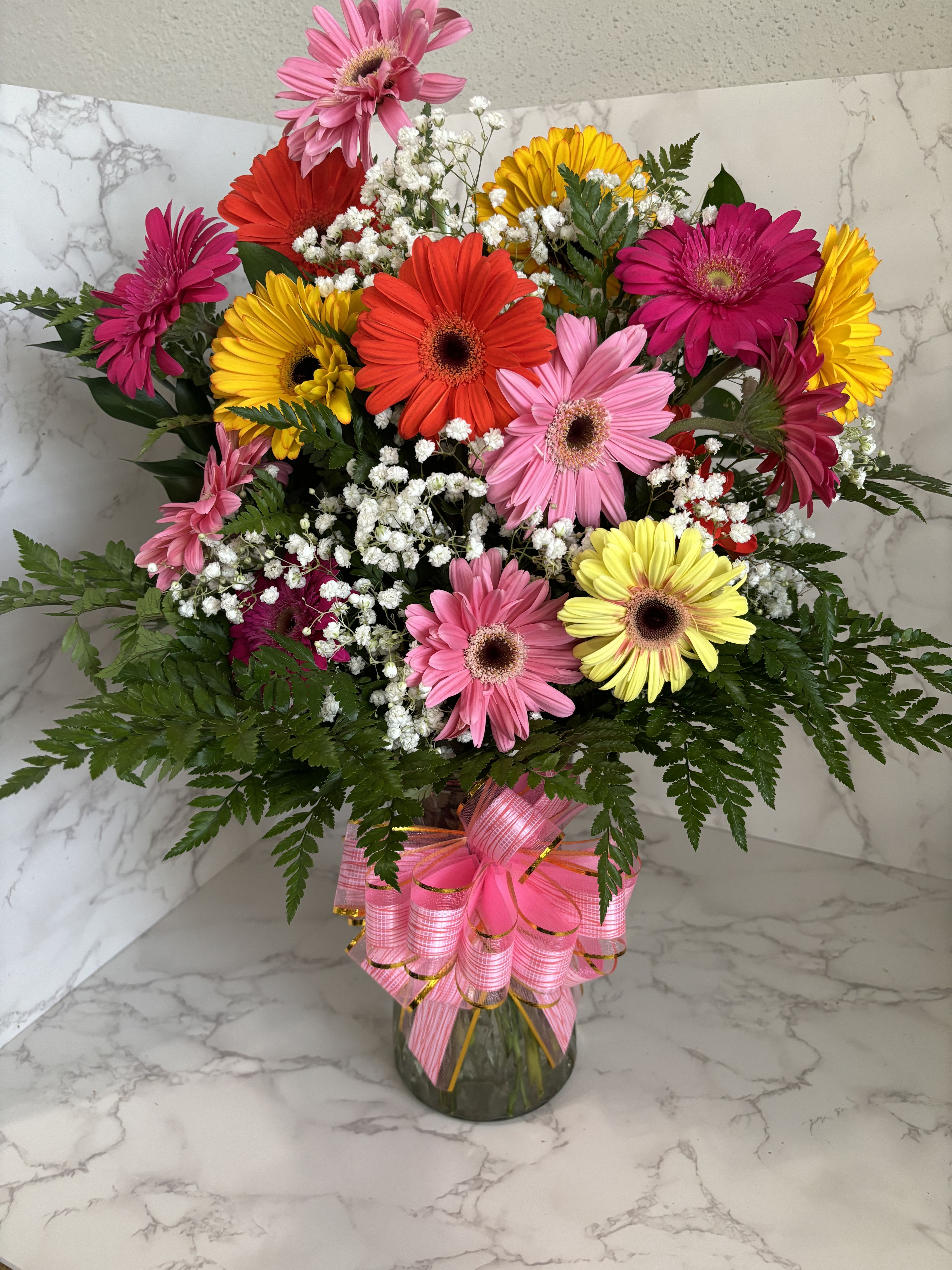 Vibrant Gerbera Daisy Bouquet in Glass Vase - Bright, cheerful, and full of life — this stunning bouquet of multicolored gerbera daisies is designed to bring joy to any occasion. Arranged with fresh greenery and delicate baby’s breath, the bold colors create a perfect balance of elegance and happiness. Presented in a clear glass vase and finished with a decorative pink bow, it makes a wonderful gift for birthdays, celebrations, or simply to brighten someone’s day.  Includes: 	•	Fresh assorted gerbera daisies in vibrant colors 	•	Baby’s breath and lush greenery accents 	•	Clear glass vase with decorative bow   Perfect for gifting, decorating, or surprising someone special. 