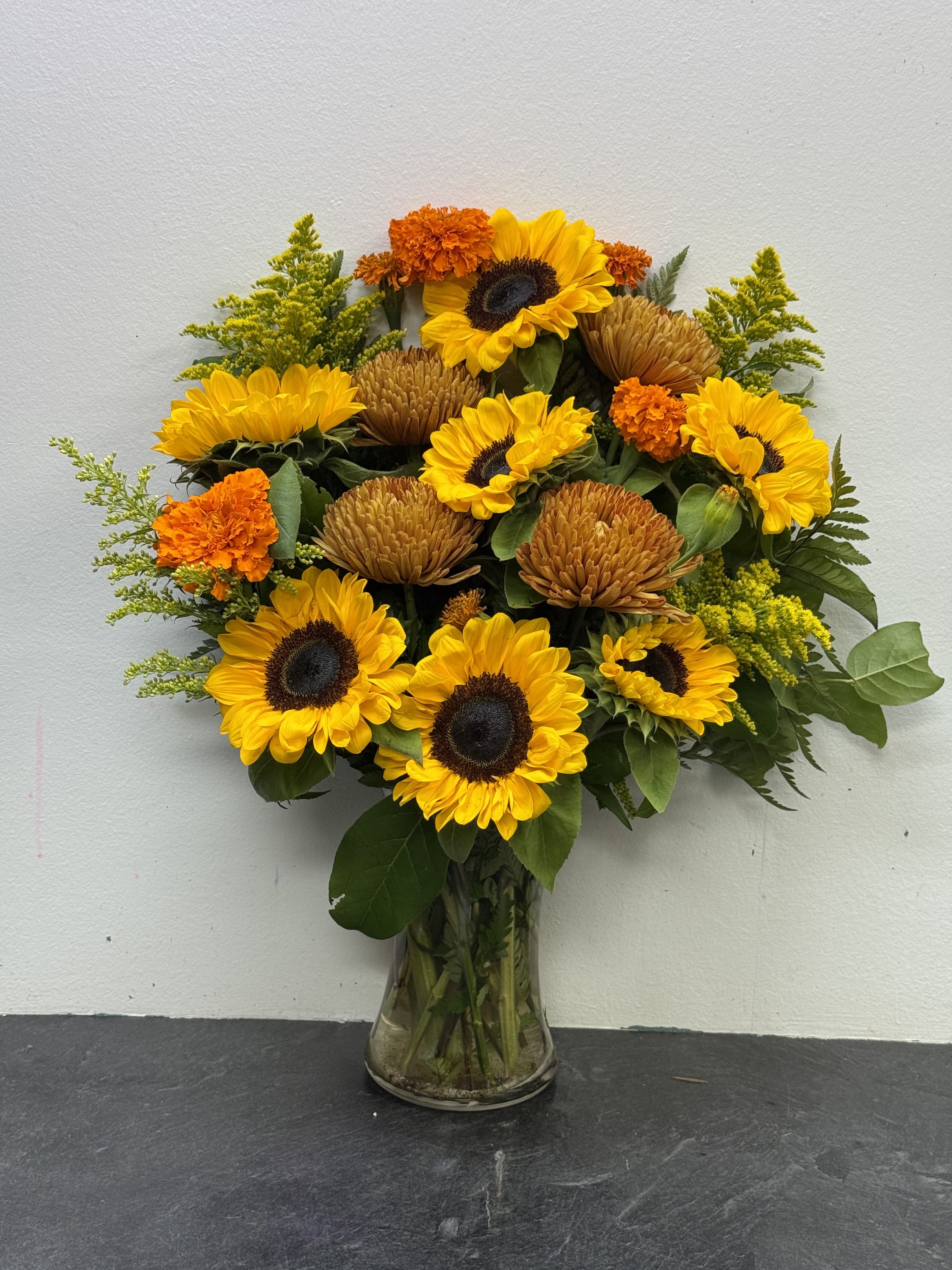 Harvest Radiance Bouquet - Bright and cheerful, this radiant arrangement captures the warmth of a sunny day. Bold sunflowers steal the spotlight, beautifully paired with golden mums, vibrant orange marigolds, and lush greenery. Hand-designed in a clear glass vase, this bouquet is perfect for bringing joy, celebrating special occasions, or adding a touch of sunshine to any space.