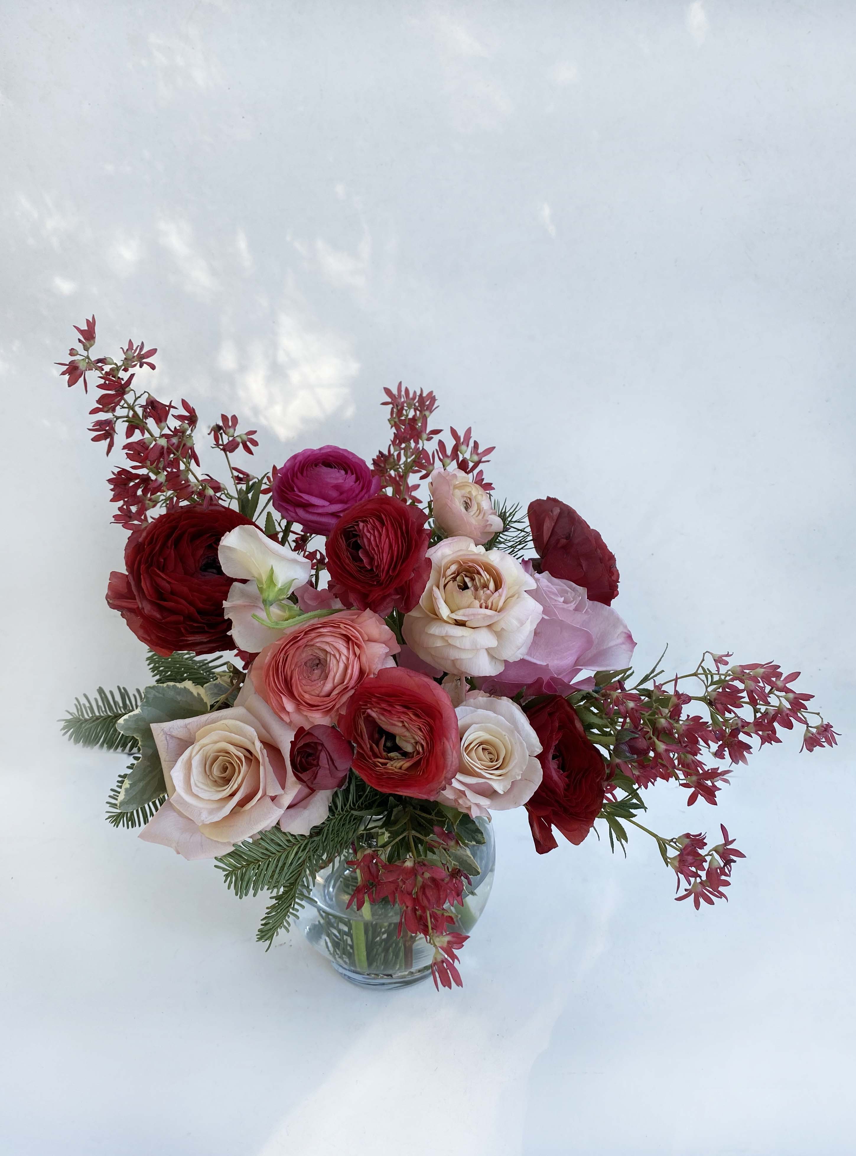 Merry arrangement - Beautiful warm blushing colors adorn this arrangement of ranunuclus, roses, and seasonal greens placed in a cute round vase, 5&quot; by 5&quot;. Perfect to send to loved ones this Christmas or place at your table.