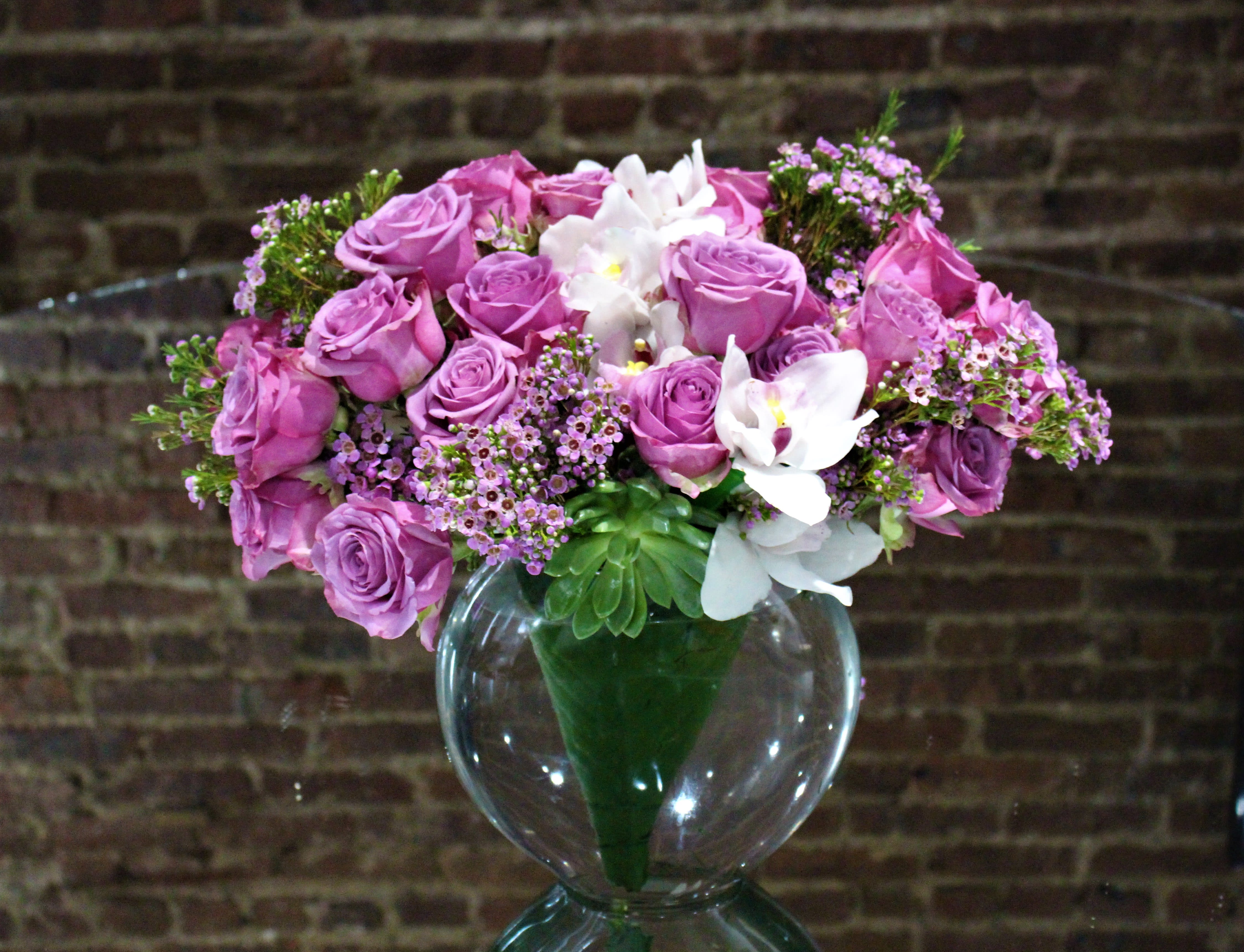 Lucious Lavender Roses with White Cymbidiums: A Louis Konstantinos Design  - An elegant display of lovely lavender roses, escorted by a trail of white cymbidium orchids accented with pink wax flower and designed in our signature eight inch pillow styled vessel.  Orientation: All-Around All prices in USD ($) Standard, Deluxe (SHOWN), Premium 