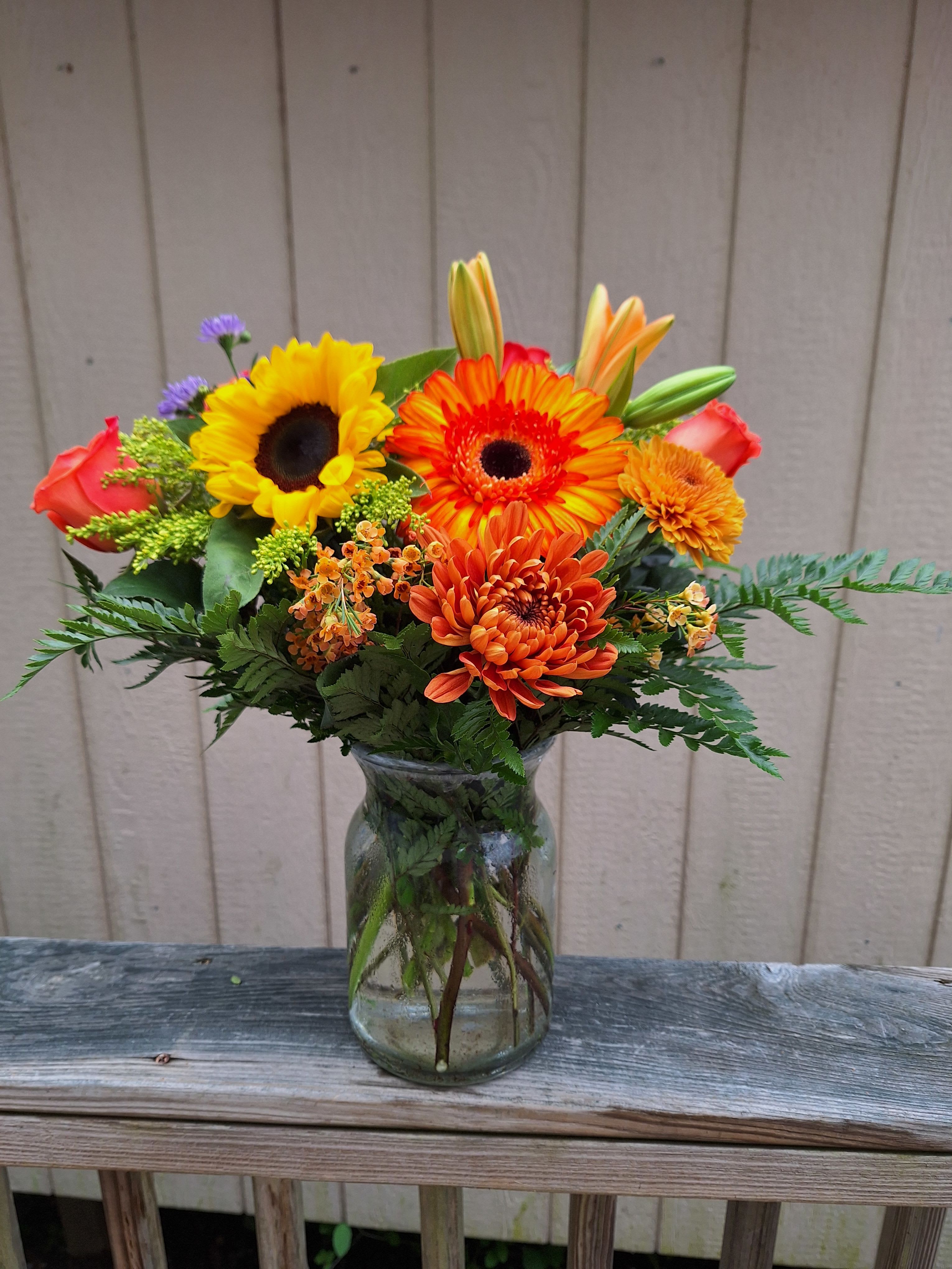 Fall Delight Bouquet - Orange gerberas, sunflowers, bronze mums, orange lilies, orange roses, orange wax flower, solidago and purple monte arranged in a milk jug vase