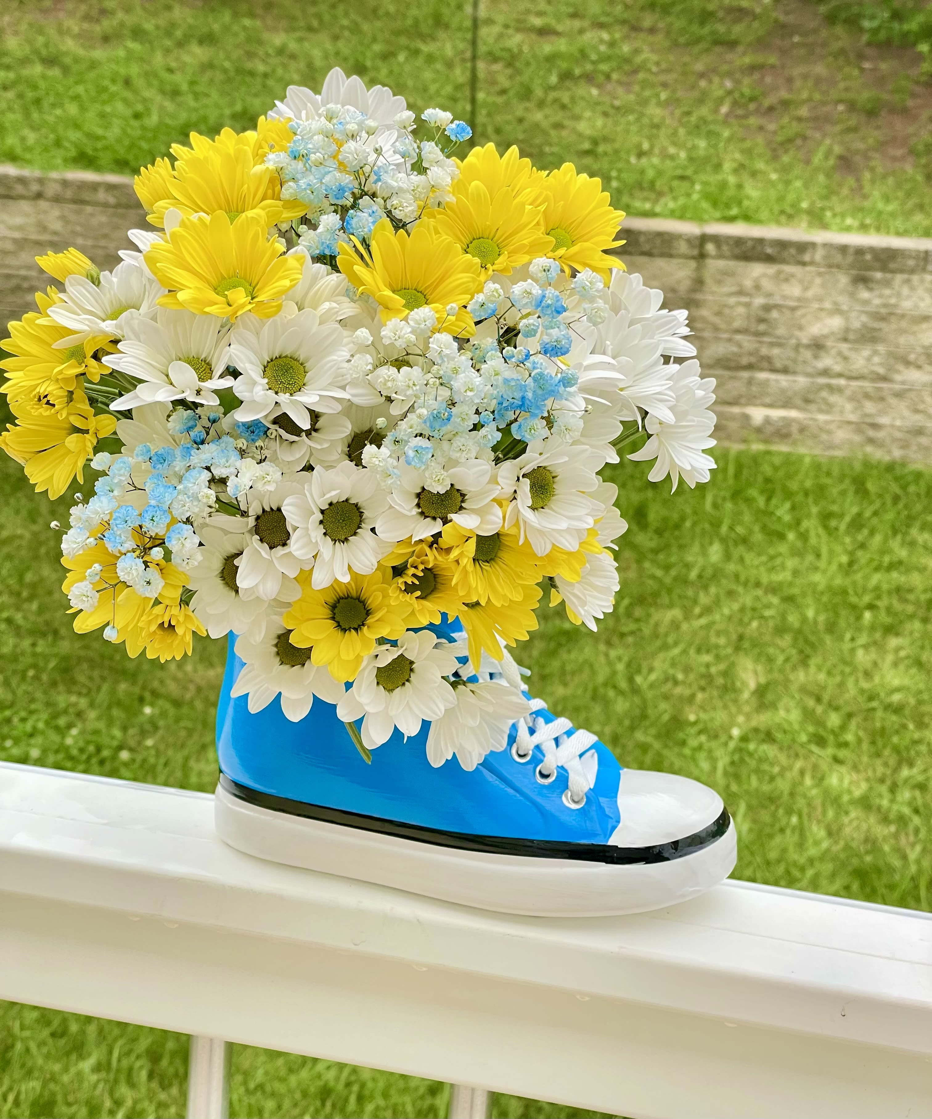 Kicks &amp; Daisies - Step up your gift-giving game with our Kickin' It With Daisies arrangement! This vibrant and playful bouquet is guaranteed to bring smiles and a fresh pop of color to any space.  We've filled a super cool ceramic high-top sneaker vase (in a brilliant blue with classic white laces!) with an abundance of cheerful blooms. You'll find a delightful mix of sunny yellow daisies and crisp white daisies, creating a classic, happy contrast. Adding a touch of whimsy and ethereal charm, delicate sprigs of light blue baby's breath are nestled among the daisies, mimicking fluffy clouds or a clear sky.  This unique arrangement is perfect for:   - Graduation gifts or celebrating new beginnings.  - A fun birthday surprise for friends, teens, or anyone young at heart!  - Brightening up a child's room or a casual living space.  - Showing school spirit with a pop of blue and yellow!  It's not just a bouquet; it's a statement piece and a keepsake that will be cherished long after the flowers are gone.  Height: 12 in Width: 8 in