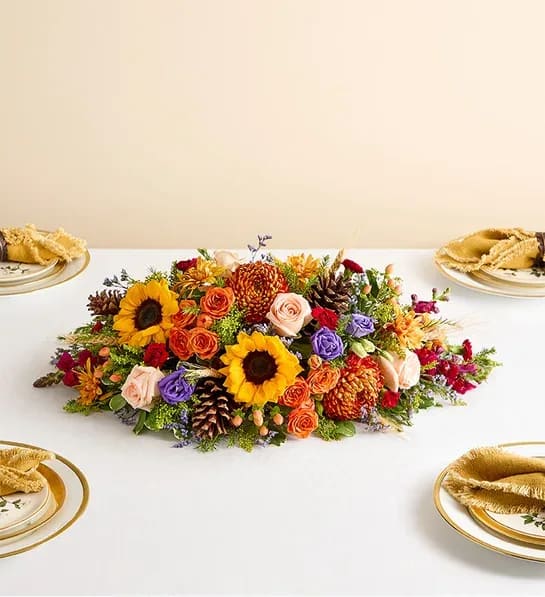 Rustic &amp; Cozy Centerpiece - NEW... This vibrant centerpiece transforms any table into the setting for a harvest celebration. Sunflowers, roses, lisianthuses, and more blooms in a dazzling array of autumnal colors make this a memorable addition to any gathering.  Large arrangement measures approximately 10.5”H x 20”L Small arrangement measures approximately 10”H x 18”L We hand-design each arrangement, so colors and varieties may vary due to local availability