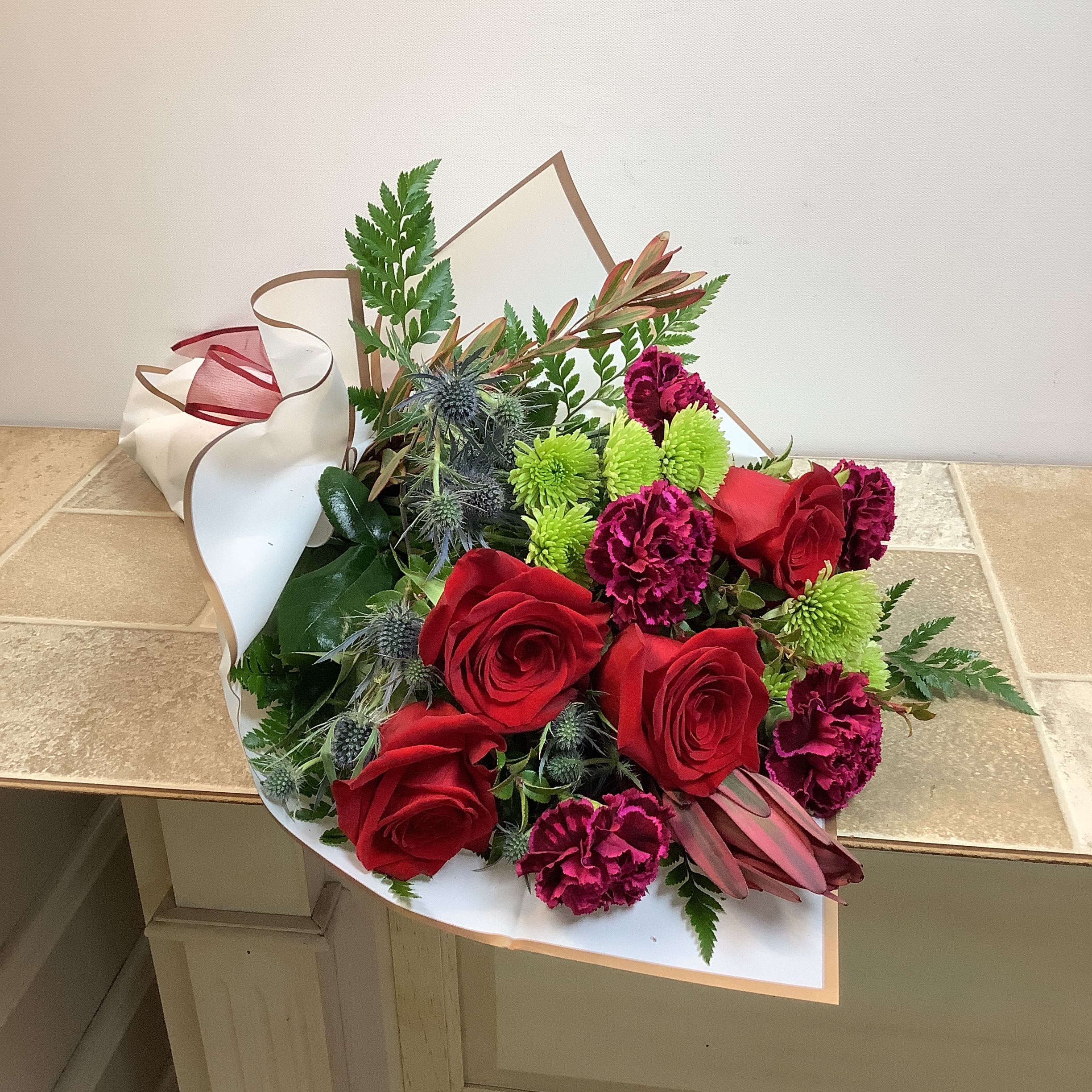 Crimson Crush - Show your love with this vibrant, wrapped flower bouquet!  Stunning red roses and ruffled magenta carnations are complemented by striking leucadendron, bright green athos mums, and textural blue thistle. A lush assortment of green foliage adds depth. A wrap with subtle gold trim and hand-tied bow offer elegant finishing touches and contemporary presentation.  Primary flowers: rose, carnation, mum, chrysanthemum