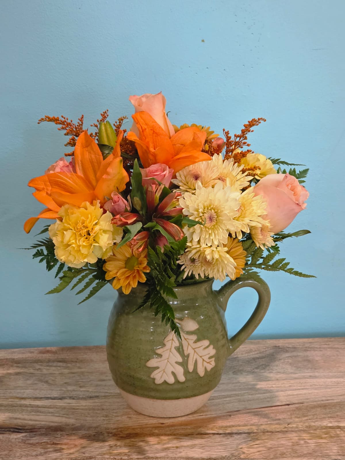 Sunset in Bloom - This charming floral arrangement bursts with warmth and joy, featuring a delightful blend of peach roses, vibrant orange lilies, cheerful orange alstroemeria, sunny yellow carnations, and playful orange daisies. Accented with fine greenery and delicate filler, each bloom is carefully arranged to create a balanced harmony of color and texture.  Presented in a rustic green ceramic pitcher with a leaf motif, this design radiates natural beauty and timeless elegance. Perfect for brightening any room, celebrating special occasions, or gifting a heartfelt gesture, it combines rustic charm with a fresh, uplifting palette that captures the spirit of joy and warmth.