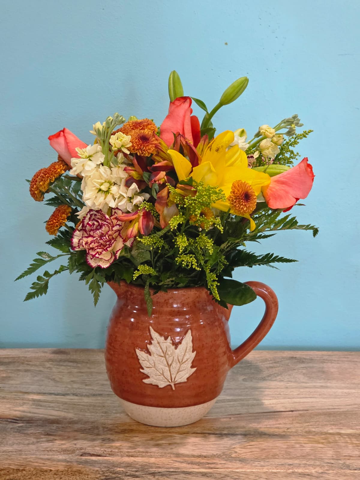 Maple Glow - Bursting with the warmth of autumn tones, this radiant arrangement features golden yellow lilies, vibrant orange roses, sunny yellow stock, cheerful button mums, and lively orange alstroemeria. Each bloom is carefully accented with lush greenery and delicate filler, creating a perfect balance of color, texture, and charm.  Designed in a rustic maple-leaf ceramic pitcher, this piece combines natural elegance with seasonal warmth. It’s an ideal choice for brightening a home, celebrating special moments, or gifting someone with a heartfelt touch of beauty that captures the essence of cozy gatherings and golden days.