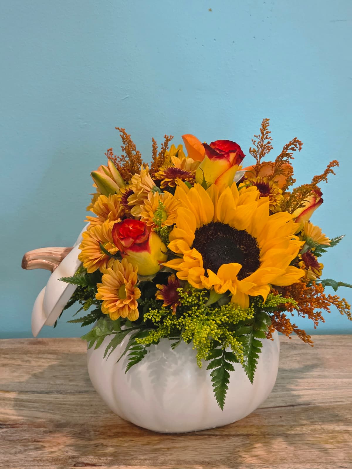 Pumpkin Glow - Celebrate the beauty of the season with this radiant design, featuring a brilliant sunflower, golden yellow roses, fiery orange lilies, cheerful orange daisies, and delicate filler blooms, all nestled among lush greenery. Each element is thoughtfully arranged to create a warm and inviting harmony of colors.  Presented in a white ceramic pumpkin vase, this charming centerpiece blends rustic harvest style with fresh elegance — making it perfect for autumn gatherings, seasonal décor, or as a heartfelt gift that captures the joy and coziness of fall.