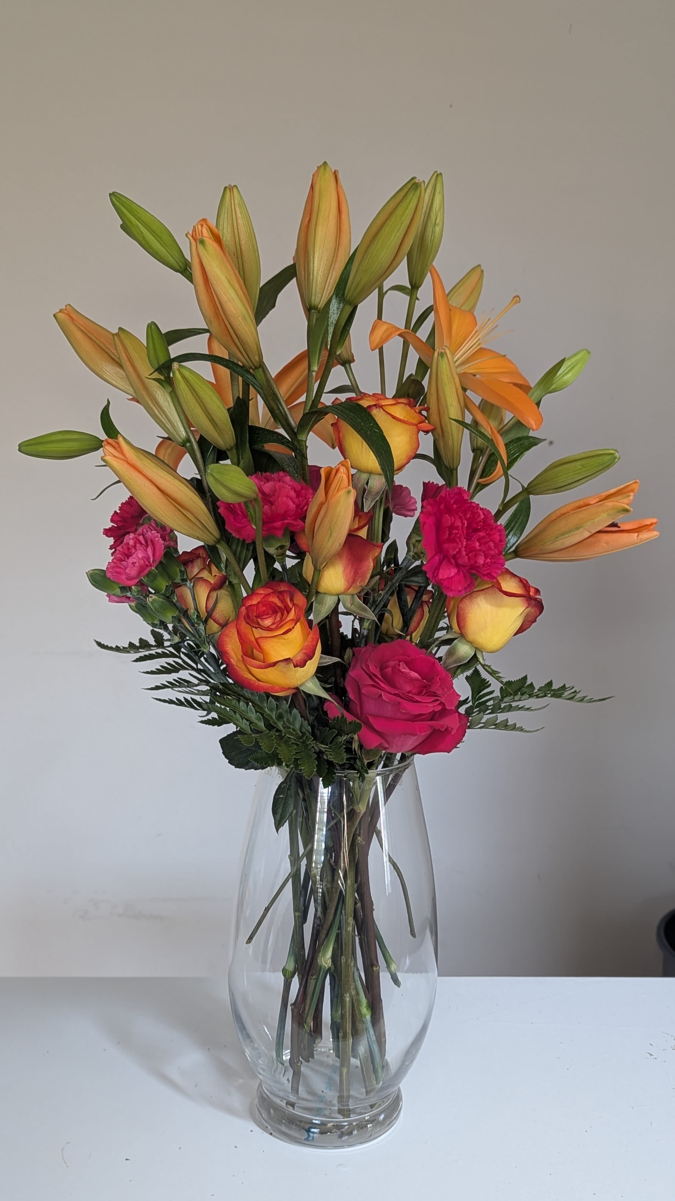 Sunset Glow  - A vivid celebration of color! Featuring fiery orange lilies, vibrant pink carnations, and two-toned roses in rich sunset hues, this tall vase design brings warmth and bold beauty to any occasion.