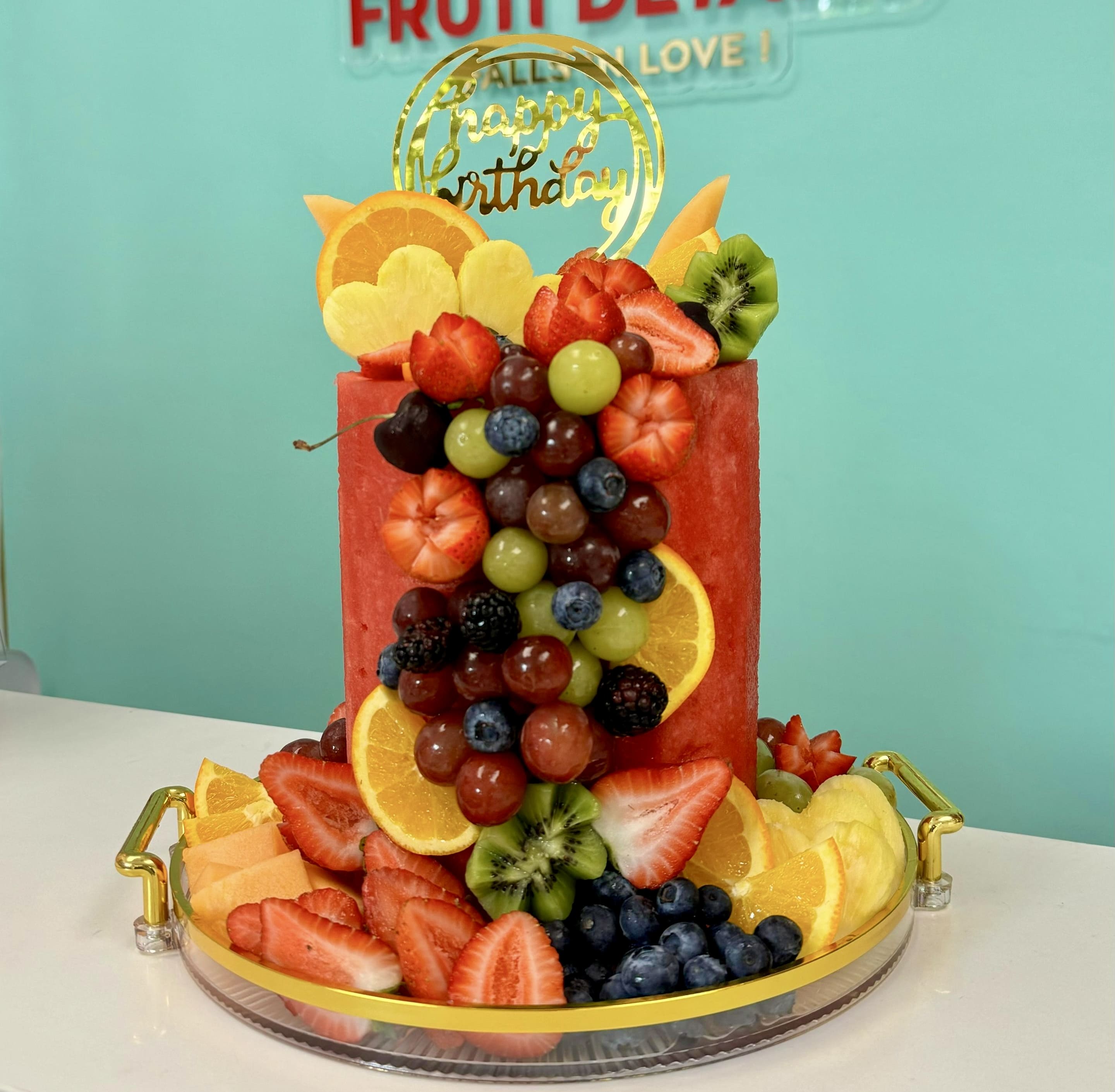 Fruit Watermelon Cake Standart -  Gifts Box Fruit Cake – Healthy Fruit Cake Birthday Buy Online the best Fruit hresh Cake, A delicious and refreshing way to celebrate! This elegant fruit “cake” is handcrafted with layers of fresh fruit and beautifully presented in a premium gift box.  This arrangement includes:   One carved watermelon base in cake shape  Decorated with strawberries, red &amp; green grapes, blackberries, blueberries, kiwi, orange slices, cantaloupe, pineapple flowers, and cherries  Topped with a golden acrylic “Happy Birthday” sign  Served on a round acrylic tray with elegant golden handles  Protected inside a clear acrylic gift box with white lid and blue ribbon – perfect for delivery or gifting