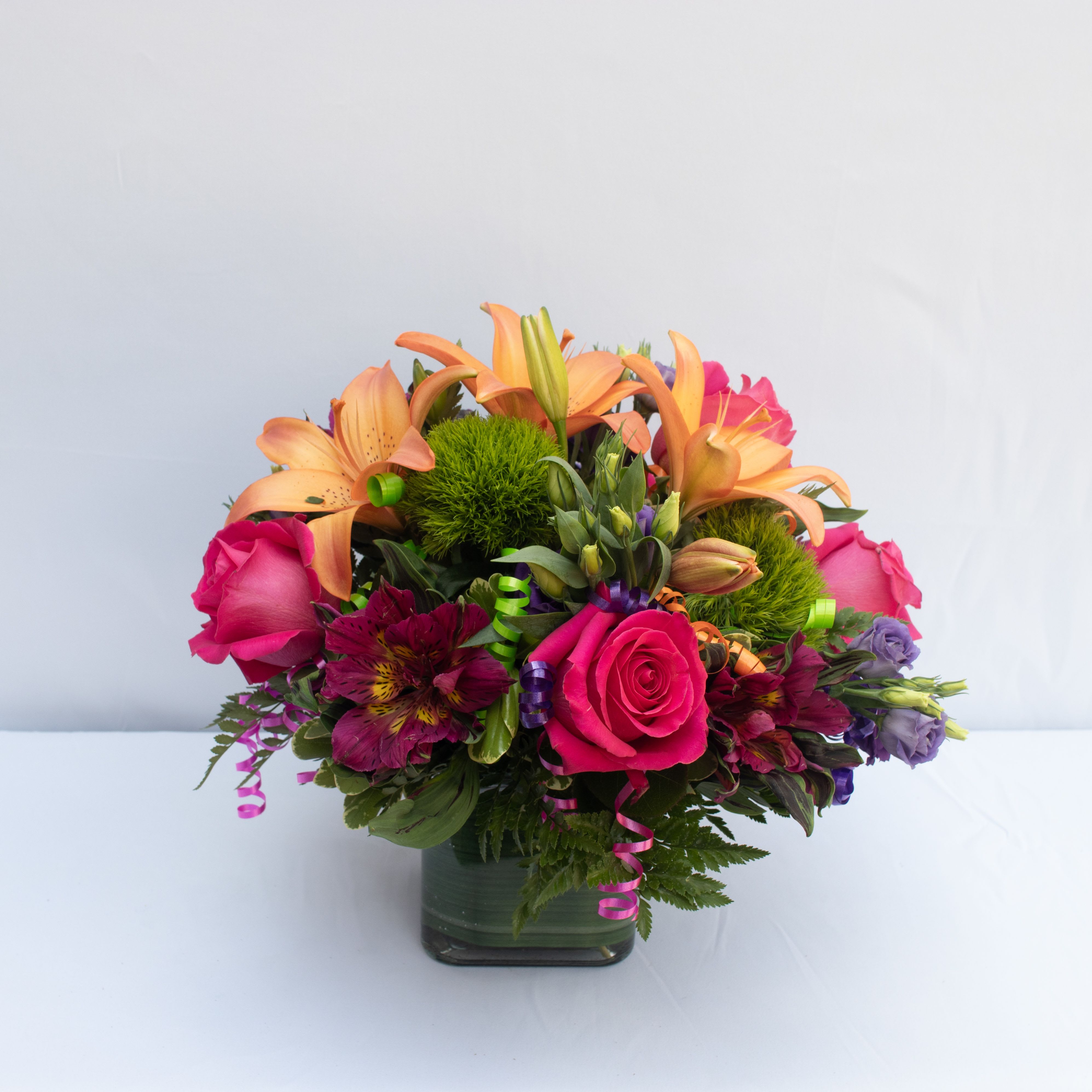 The Birthday Cheer Bouquet The Birthday Cheer Bouquet - Birthday blooms that are ready to get your recipient's special day started this flower bouquet is bright happy and ready to celebrate! Hot pink roses and orange Asiatic Lilies are vibrant and fun surrounded by purple Peruvian Lilies hot pink mini carnations green button poms purple statice and an assortment of lush greens. Accented with assorted curling ribbons to give it that party feel and presented in a clear glass cubed vase lined with ti leaf green material for added beauty this unforgettable birthday bouquet is that ultimate surprise that will make them feel the love on their big day.
