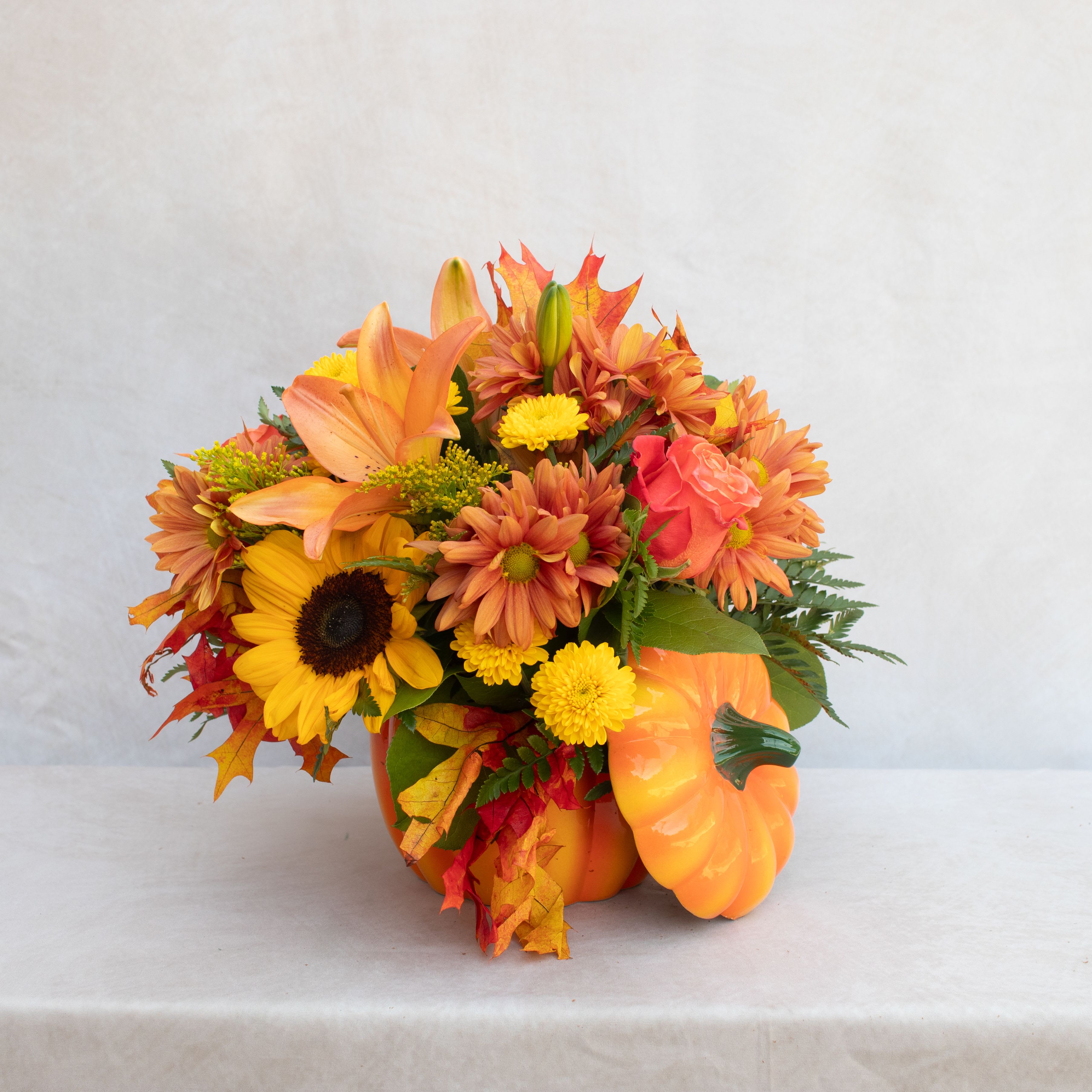 Pumpkin Spice  - A fall assortment of orange, yellows, and rust flowers like lilies, roses, sunflowers, mum, fillers, and greenery in a ceramic pumpkin.    The arrangement designed is:  10wx10h standard 12wx12h deluxe  14wx14h premium 