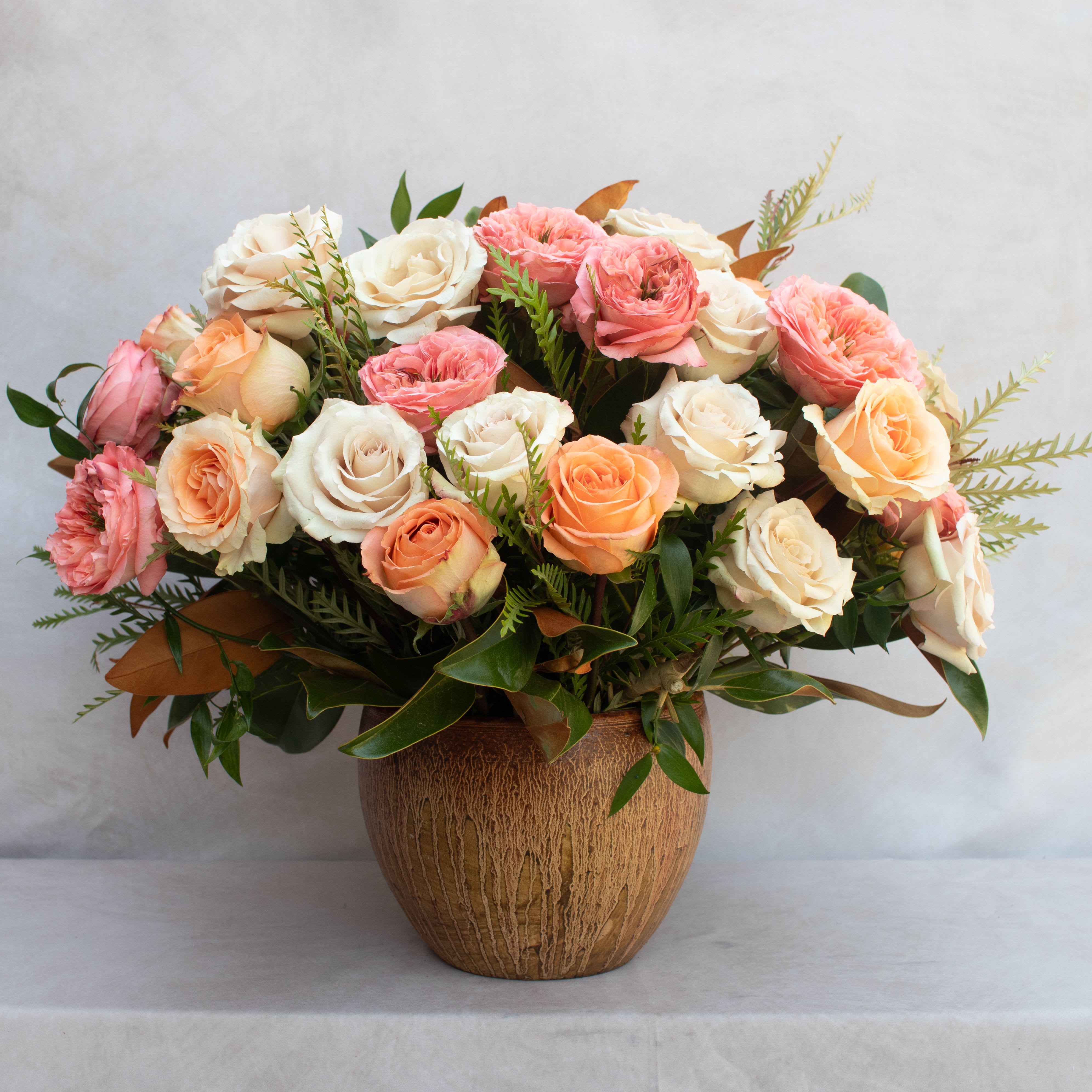 FALL FANTASY  - BOUQUET OF MIXED ROSES IN A GLAZED TERRACOTTA CONTAINER.  ARRAY OF ROSES IN COLORS AVAILABLE LIKE PEACHES, ORANGES, PINKS, WHITES, CREAMS, BEIGES, CORALS ACCENTED WITH UNIQUE GREENERY LIKE MAGNOLIA LEAVES, EUCALYPTUS, RUSCUS. ARRANGEMENT IS ABOUT 18X18 DESIGNED.  