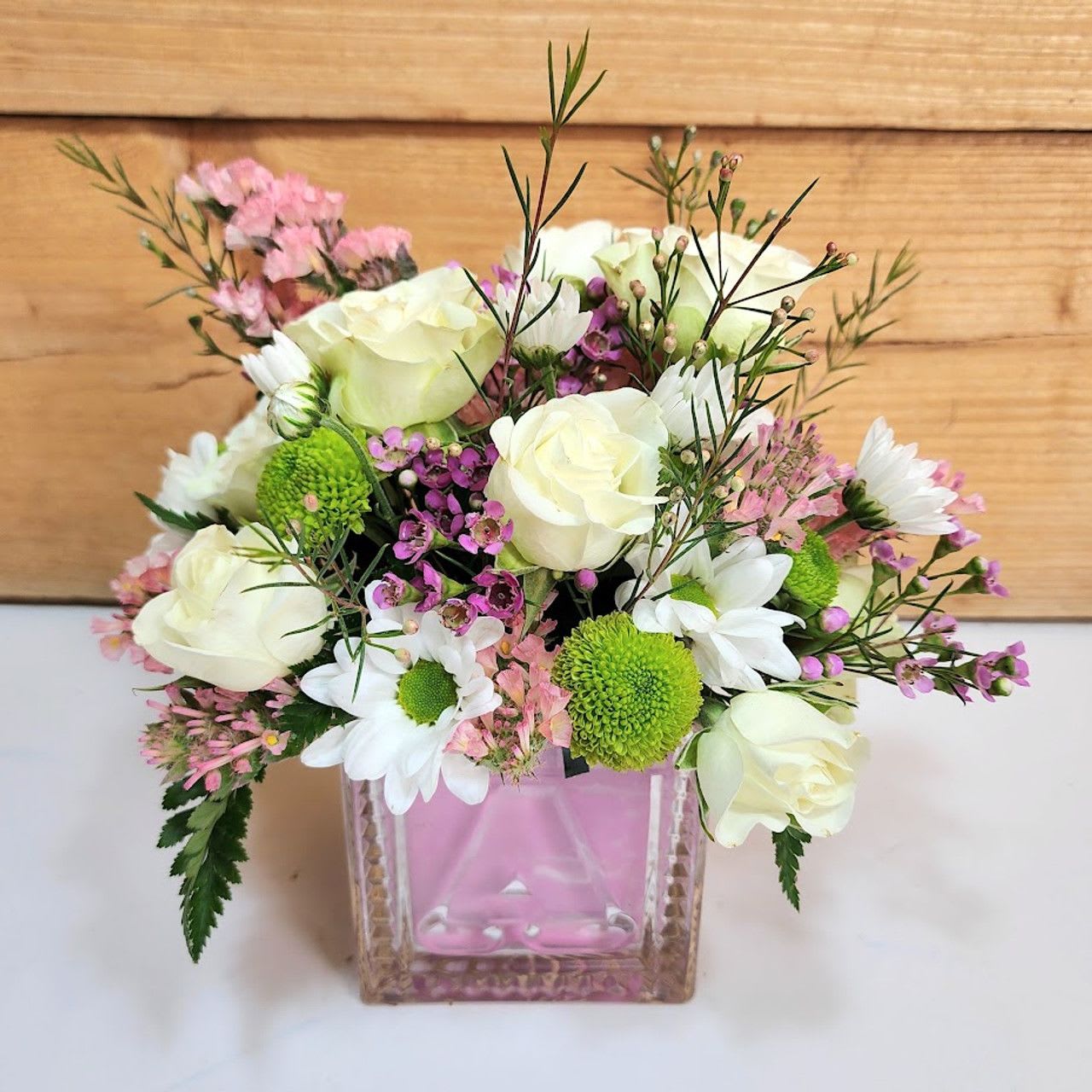 Pretty in Pink Blooms - Celebrate the joy of a new baby girl with this delightful arrangement bursting with charm and tenderness. Soft white roses, cheerful daisies, and pops of bright green mums mingle with delicate shades of pink waxflower and statice, all beautifully arranged in a keepsake pink cube vase. Perfectly sized for a nursery, hospital room, or home, this arrangement is a fresh and thoughtful way to welcome her into the world and congratulate the proud parents.