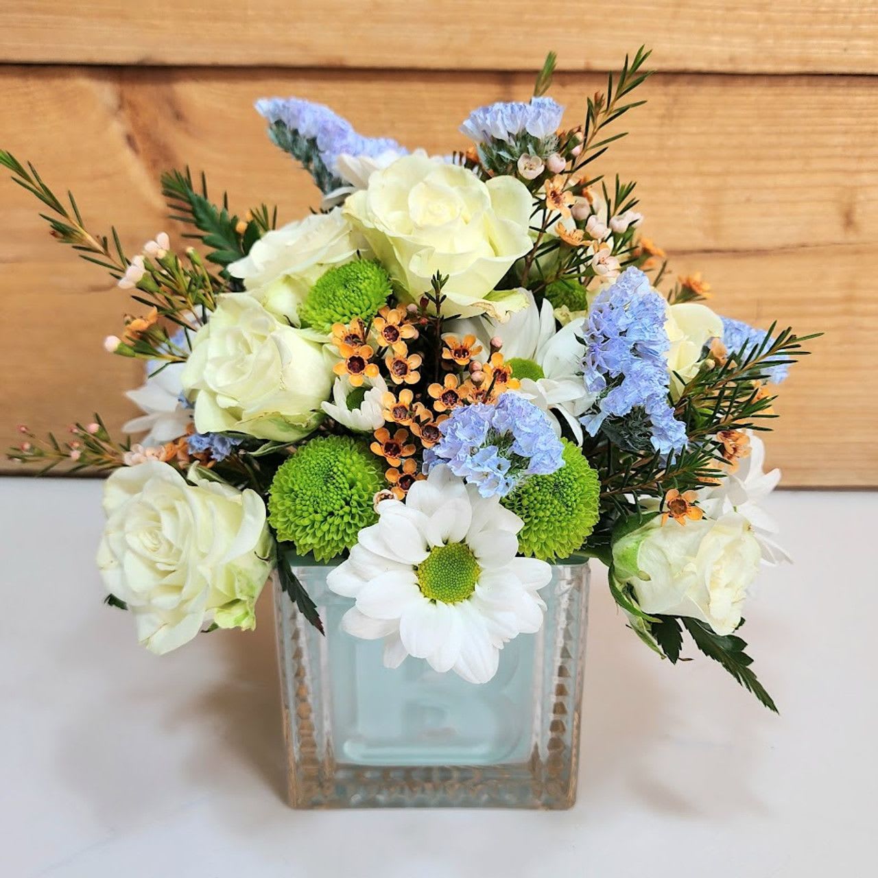 Handsome in Blue Blooms - Celebrate the arrival of a sweet baby boy with this charming arrangement full of fresh cheer. Creamy white roses, playful daisies, and vibrant green mums are accented with touches of soft blue statice and peach waxflower, all arranged in a keepsake blue cube vase. The soothing blend of colors makes it a perfect gift for a nursery, hospital room, or the family’s home—bringing joy and warm congratulations to the proud parents.