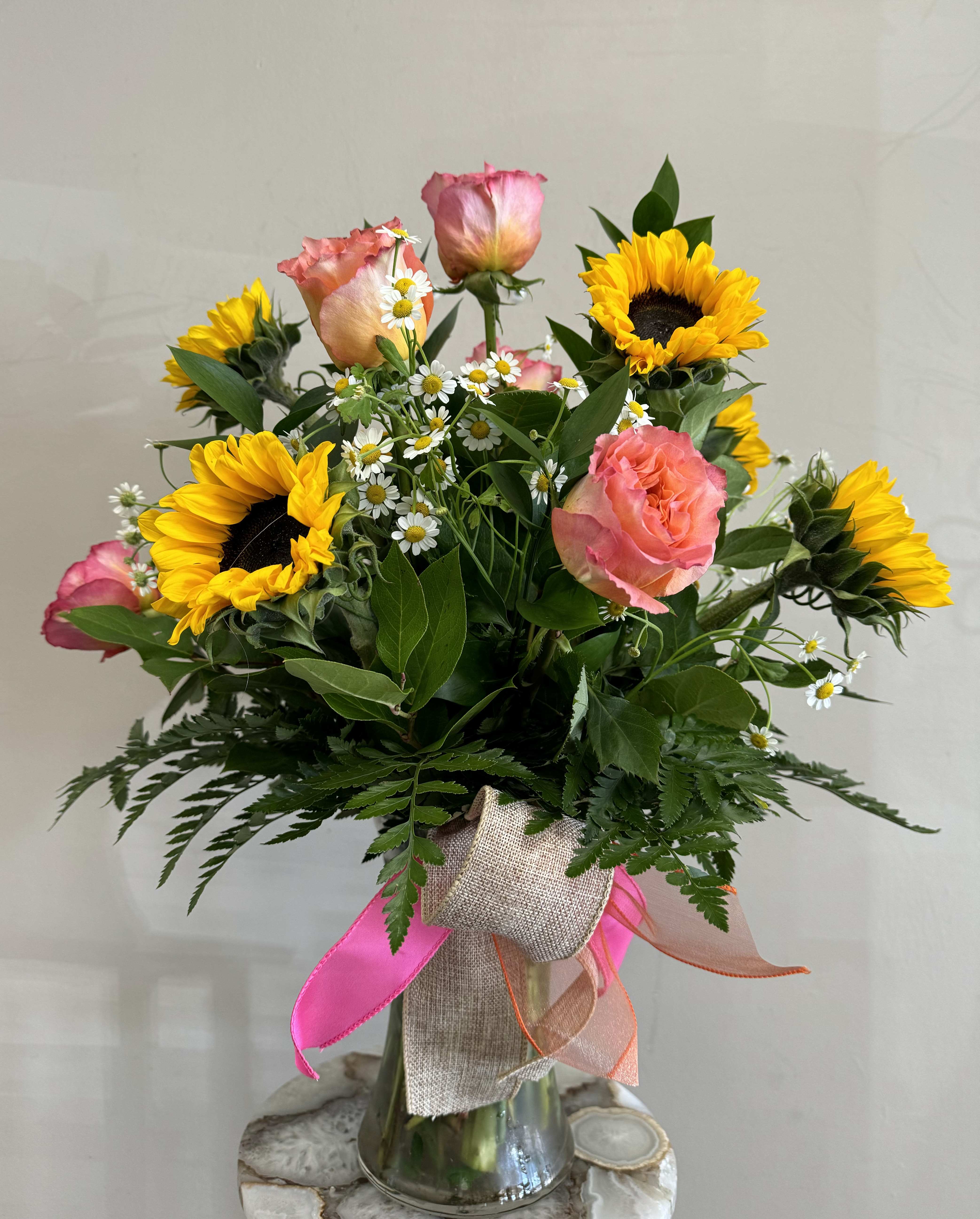 Sunburst Harmony Sunburst Harmony - In this arrangement, the warmth of the sun is captured through the bold and radiant presence of six sunflowers, their golden petals reaching out like beams of sunshine. Nestled amongst them are the fiery blooms of six orange Free Spirit roses, adding a captivating burst of color and fragrance.