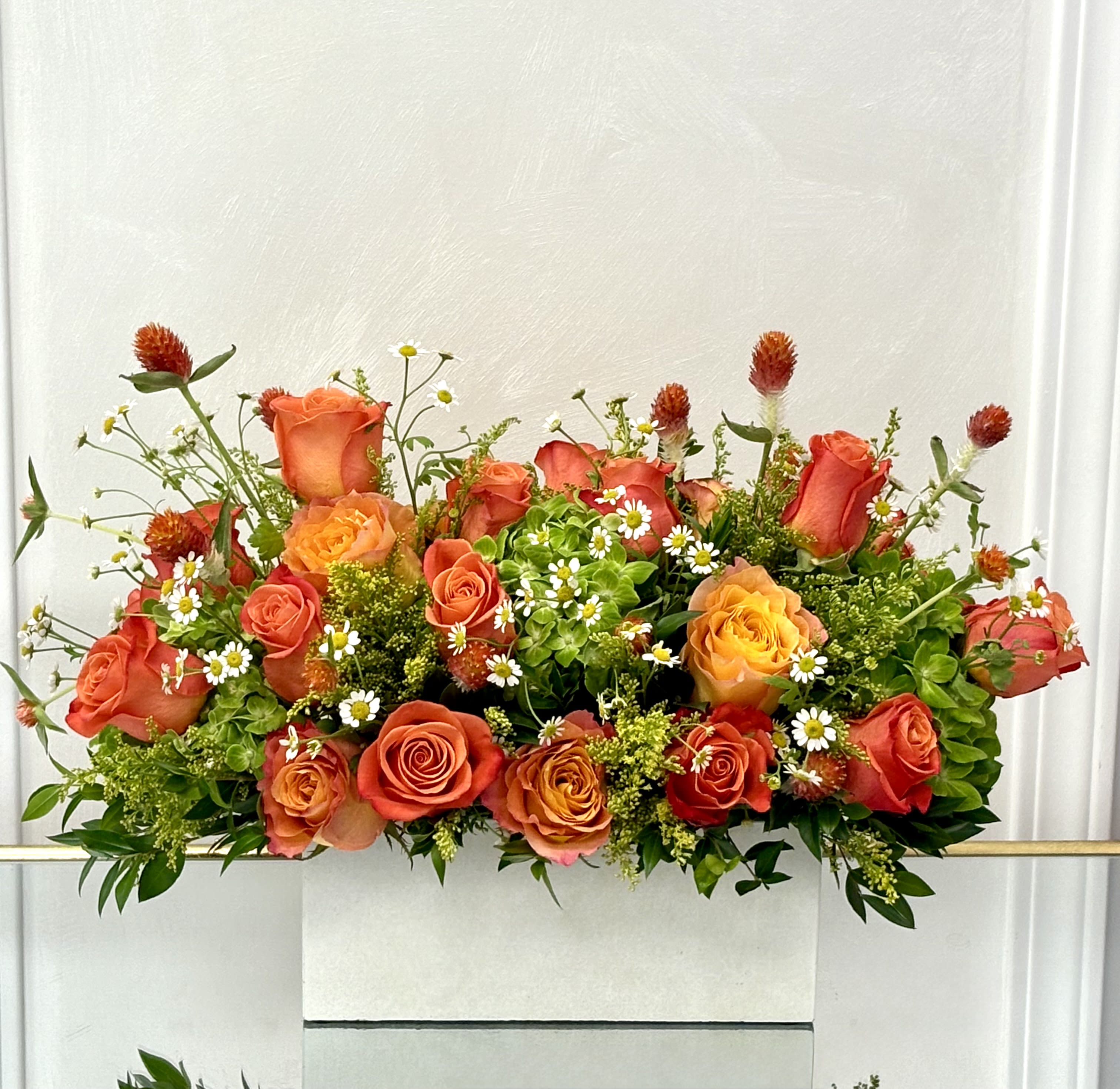 Firelight Garden By VLM Flowers - A radiant mix of orange and golden roses, accented with playful chamomile blooms, lush greenery, and touches of red gomphrena. This arrangement bursts with warmth and cheer, perfect for brightening any space or occasion.