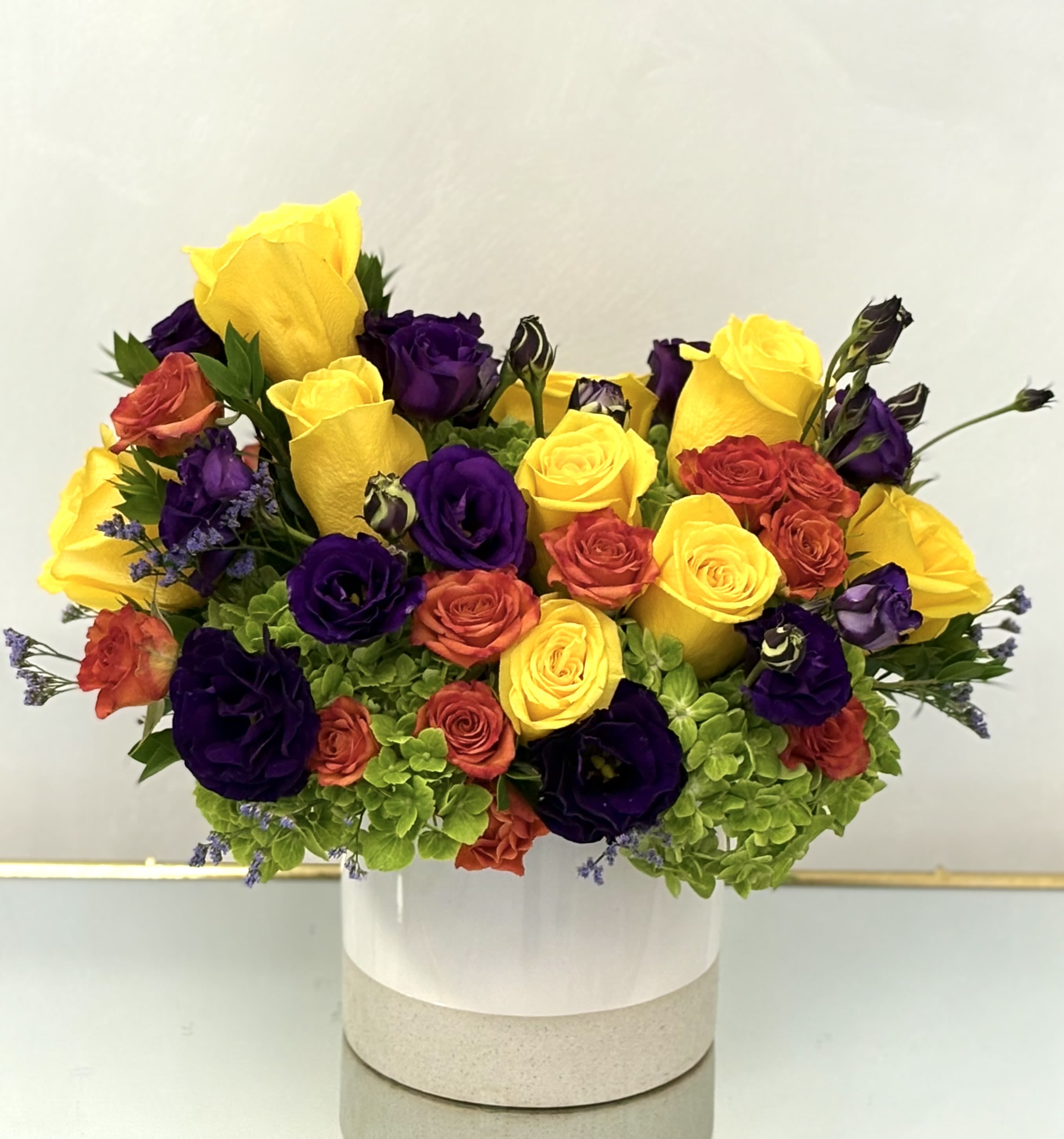Sunset Radiance By VLM Flowers - A bold, joyful burst of color that captures the warmth of a golden sunset. This luxurious arrangement features bright yellow roses, deep violet lisianthus, cheerful orange spray roses, and soft green hydrangeas. Artfully arranged in a modern white ceramic vase, it’s the perfect way to celebrate life, express gratitude, or send a radiant message of encouragement and joy.