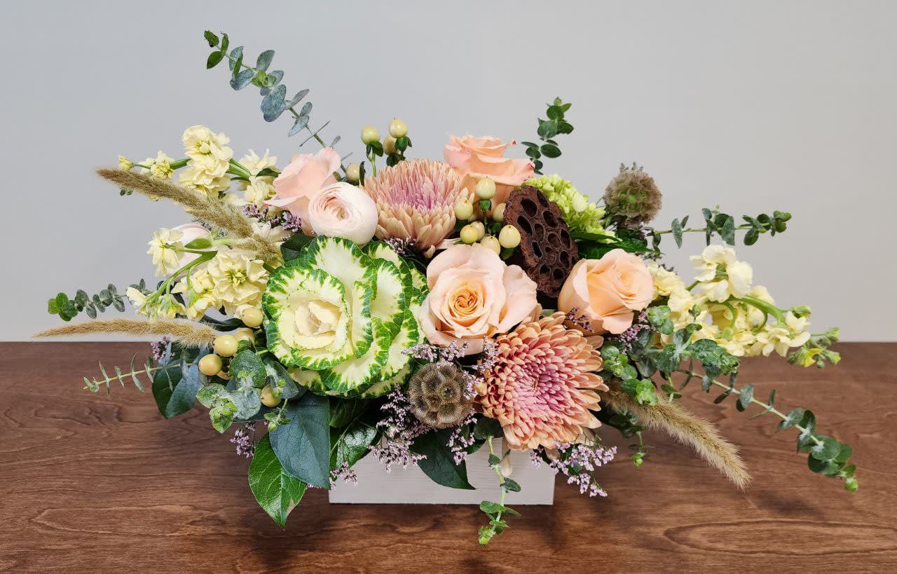Modern Simplicity - This soft and sweet centerpiece is all you need to illuminate your table setting this Thanksgiving! This combination of stockflower, roses, ranunculus, kale, and other accents comes in a white, wooden rectangular container. 
