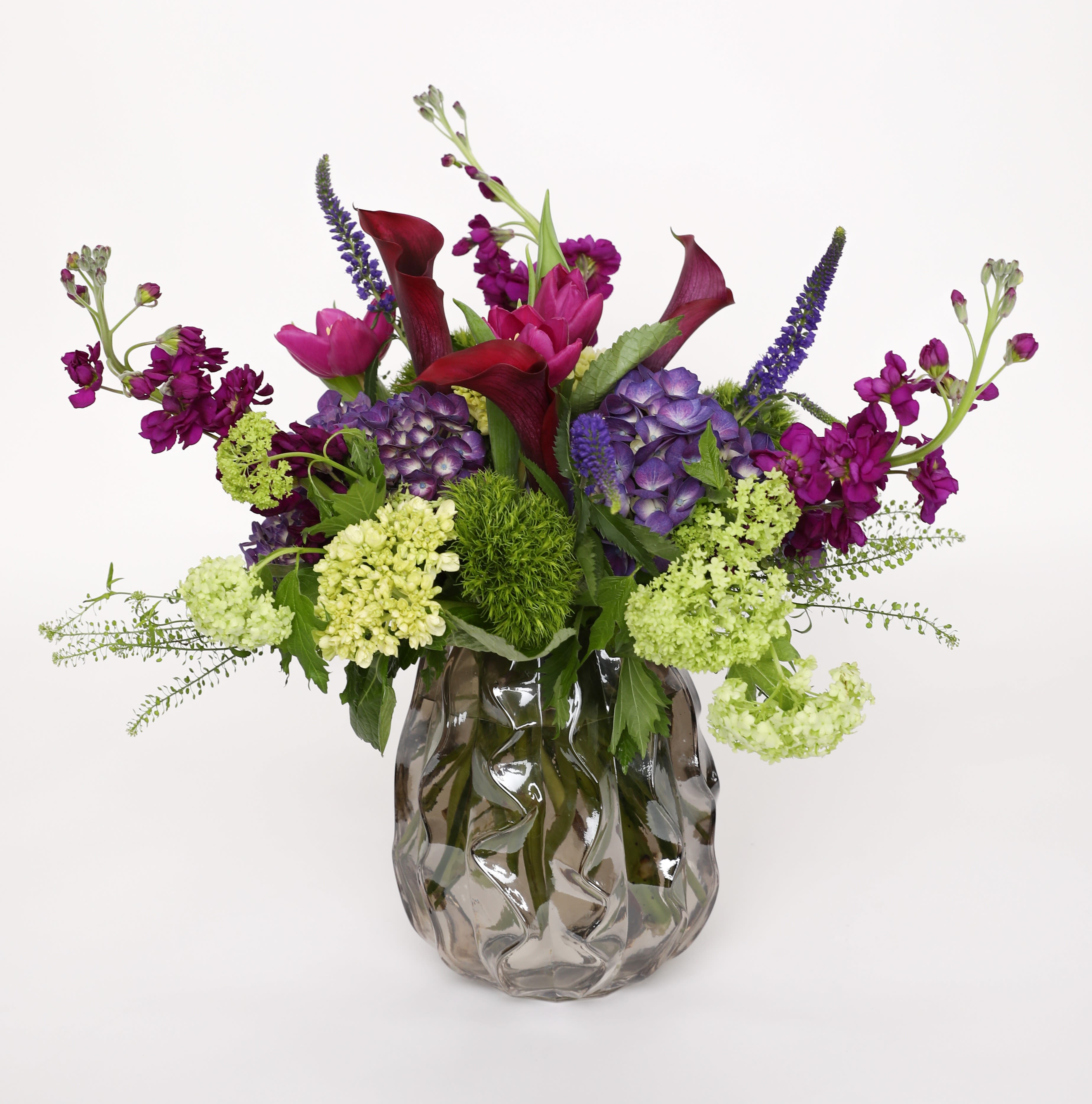 AMETHYST AMETHYST - We love everything about this all purple and green arrangement, starting with the glass vase. All purple hydrangea, mini calla lilies, stock, tulips, and veronica is surrounded by green mini hydrangea, dianthus and viburnum.