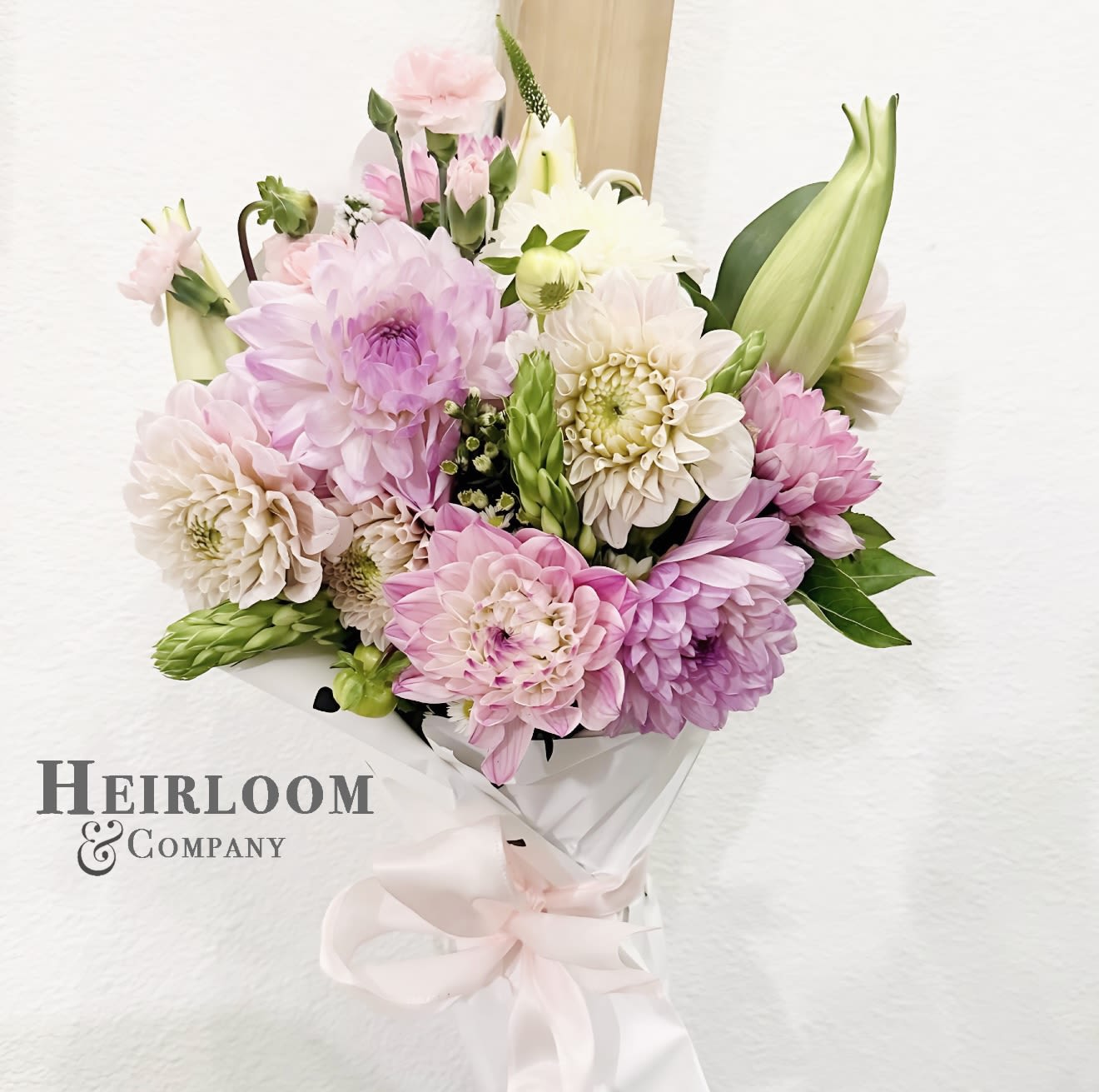 Blooming Dahlia handheld bouquet - Soft, dreamy, and oh-so romantic, this hand-tied bouquet features layers of blush and lilac dahlias, delicate spray roses, and fresh greenery wrapped in our signature heart-print paper. Tied with a blush satin bow, it’s the perfect gift to say “thinking of you” or “love you endlessly.” A chic and feminine piece that makes everyday moments feel like a love story. 