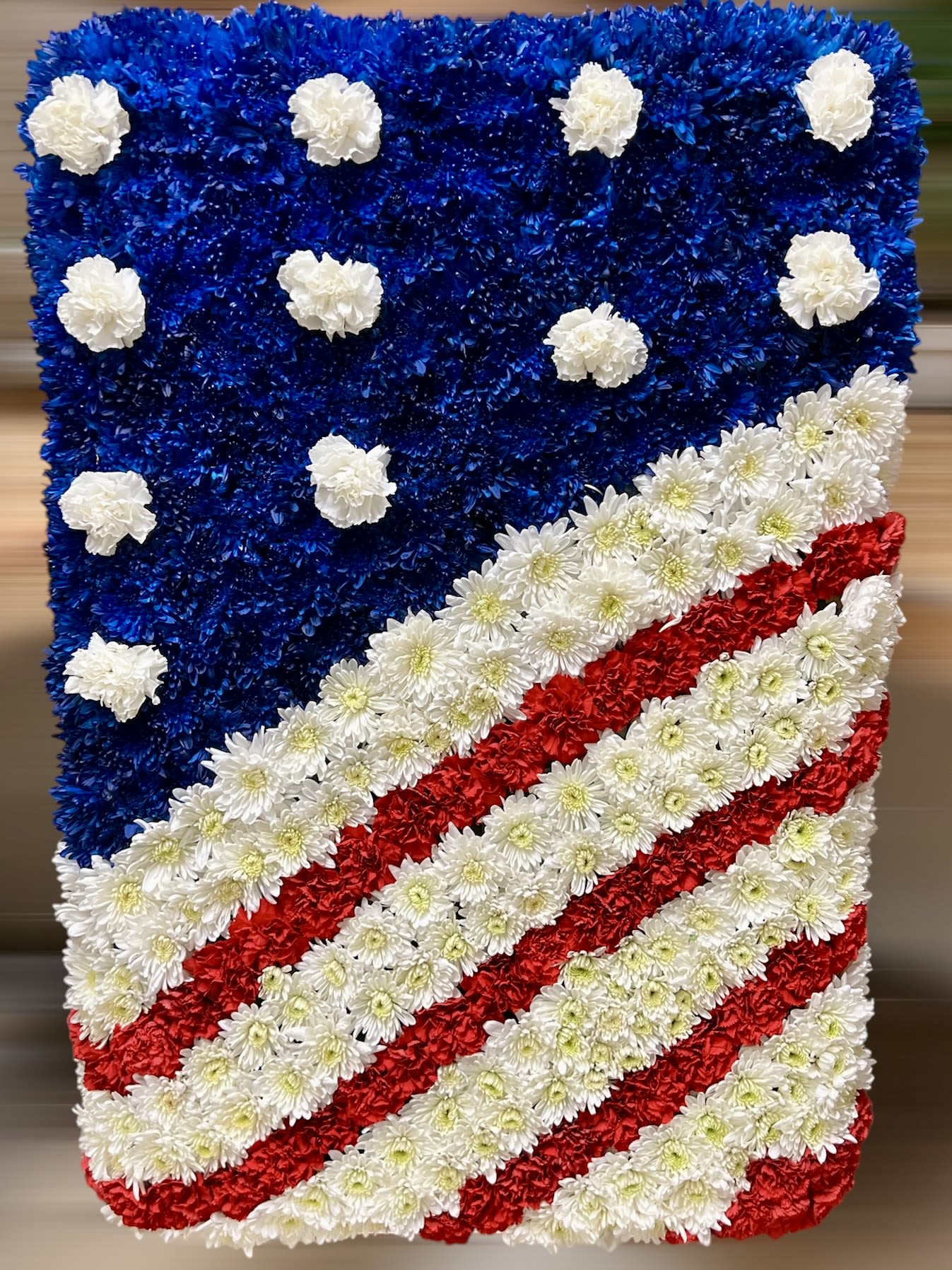 American Flag - This stunning floral arrangement is a beautiful tribute to the American flag, crafted with precision and artistry. The top left corner is filled with deep blue flowers, adorned with white blooms to represent stars, while the lower section showcases bold red and white stripes made of fresh carnations and chrysanthemums. The vibrant colors and intricate design make  This stunning floral arrangement beautifully captures the essence of patriotism, with a design inspired by the American flag. The blue background is adorned with white stars, while the vibrant red and white stripes flow diagonally, all made from carefully arranged chrysanthemums. A striking and unique tribute, perfect for national holidays, memorial events, or to express pride and unity. This floral creation is a masterpiece that combines creativity, color, and symbolism in one stunning display!  FLAG SIZE 25 X 36 Inches