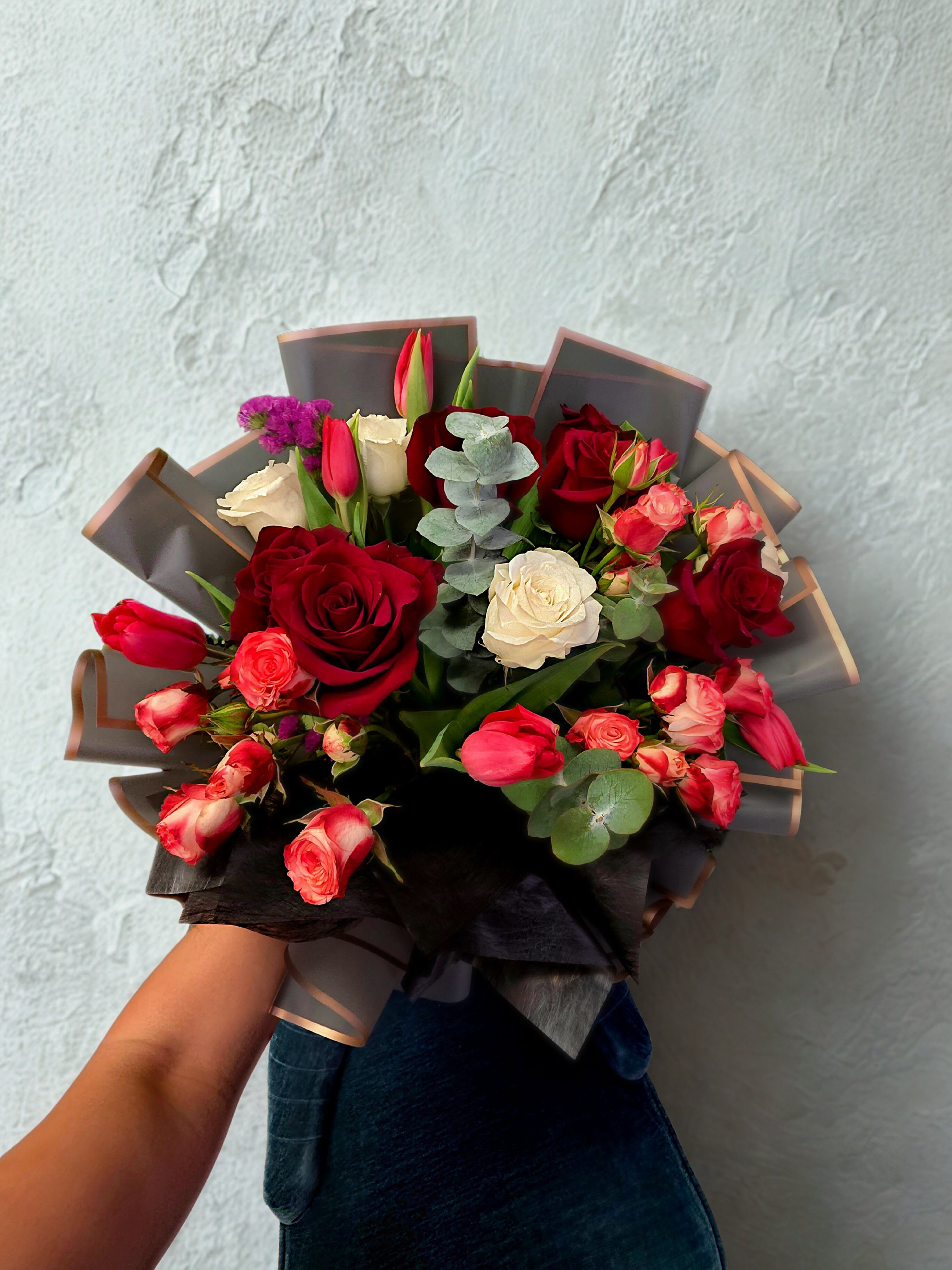 Crimson Whisper Bouquet Crimson Whisper Bouquet - Passionate, elegant, and timeless — the Crimson Whisper Bouquet combines the richness of deep red roses, the softness of cream blooms, and the charm of pink spray roses and tulips, all nestled with fresh eucalyptus leaves for a touch of modern freshness. Wrapped in sleek black and nude Korean-style paper, this bouquet speaks the language of love and sophistication. Perfect for anniversaries, romantic surprises, or any heartfelt gesture. Each piece is handcrafted with care, making it a stunning expression of emotion — bold yet refined, classic yet modern.