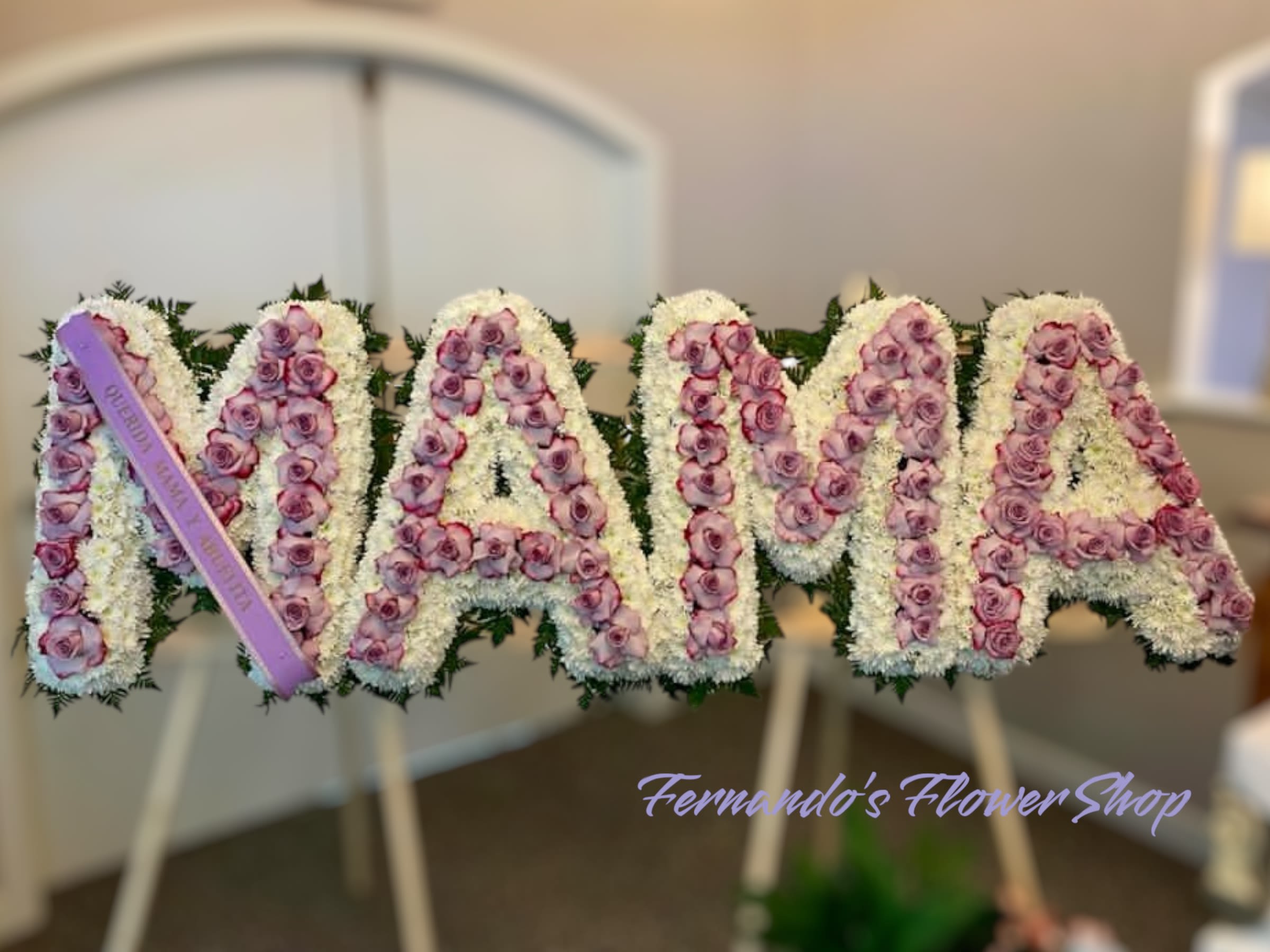 MAMA - This is a beautiful floral tribute spelling out “MAMA,” symbolizing love, honor, and remembrance. Each letter is crafted with a base of white chrysanthemums, representing purity and peace. Soft lavender roses fill the letters, symbolizing admiration, grace, and everlasting love. A delicate lavender ribbon with an inscription adds a personal touch. This heartfelt arrangement is perfect for honoring a beloved mother’s memory with elegance and affection.  LETTER HIGH 22 INCHES COLOR CAN BE CHANGED AT NO EXTRA CHARGE