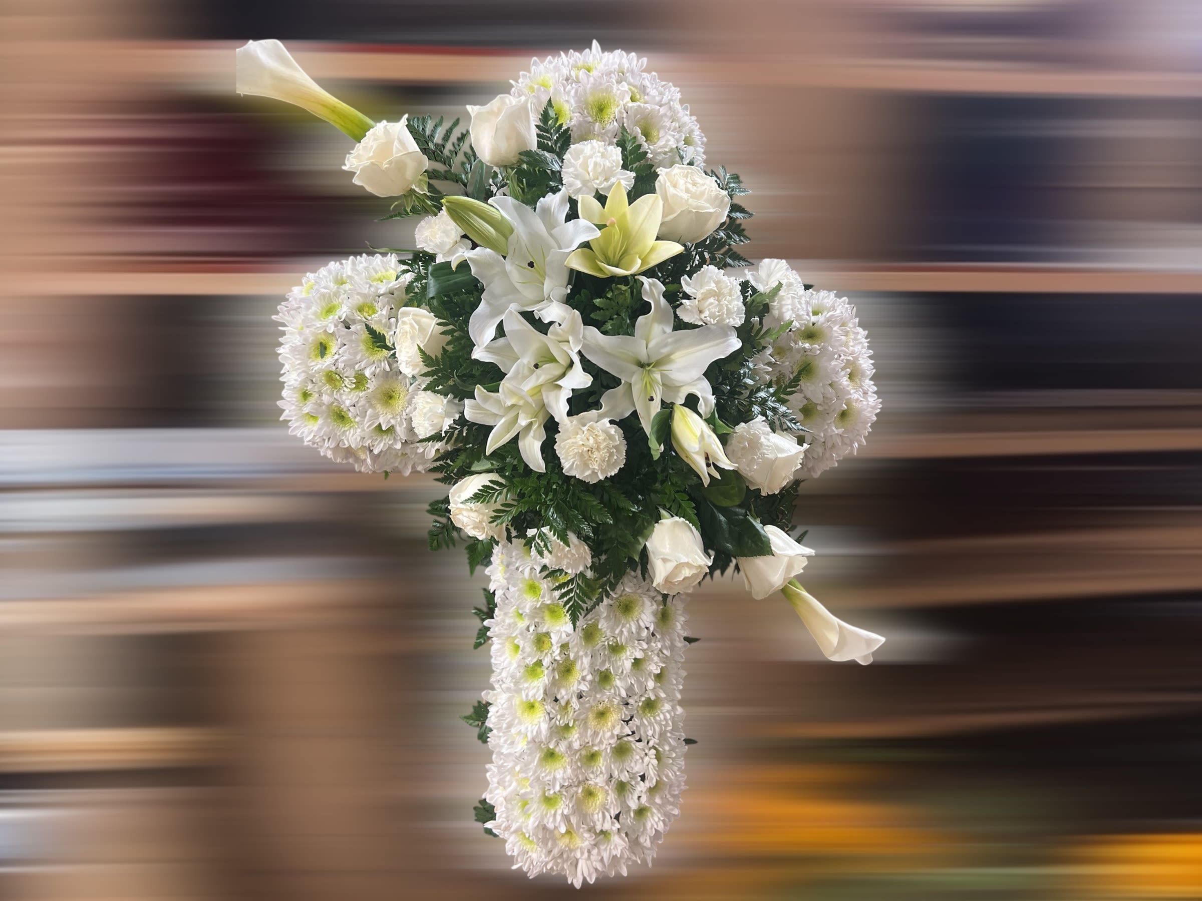 Peacefully Resting 2 - This beautiful floral cross arrangement is a timeless expression of peace and faith. Designed with pure white chrysanthemums as the base, it symbolizes purity and serenity. At the center, elegant white lilies and roses are delicately arranged, adding sophistication and depth. Accents of lush greenery complete the design, creating a harmonious and natural look. This stunning piece is perfect for honoring a loved one at a memorial or funeral service, offering a heartfelt message of sympathy and remembrance.
