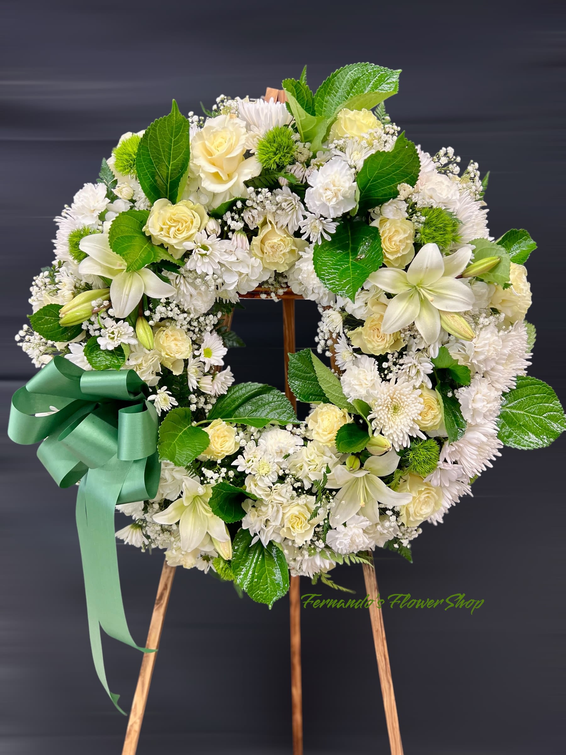 Serene &amp; Elegant - This is a circular funeral wreath featuring a mix of white and pale yellow roses, lilies, chrysanthemums, and baby’s breath. It is accented with lush green leaves and a large green ribbon, creating a serene and elegant tribute. The wreath is displayed on a wooden easel, with “Fernando’s Flower Shop” subtly marked at the bottom.