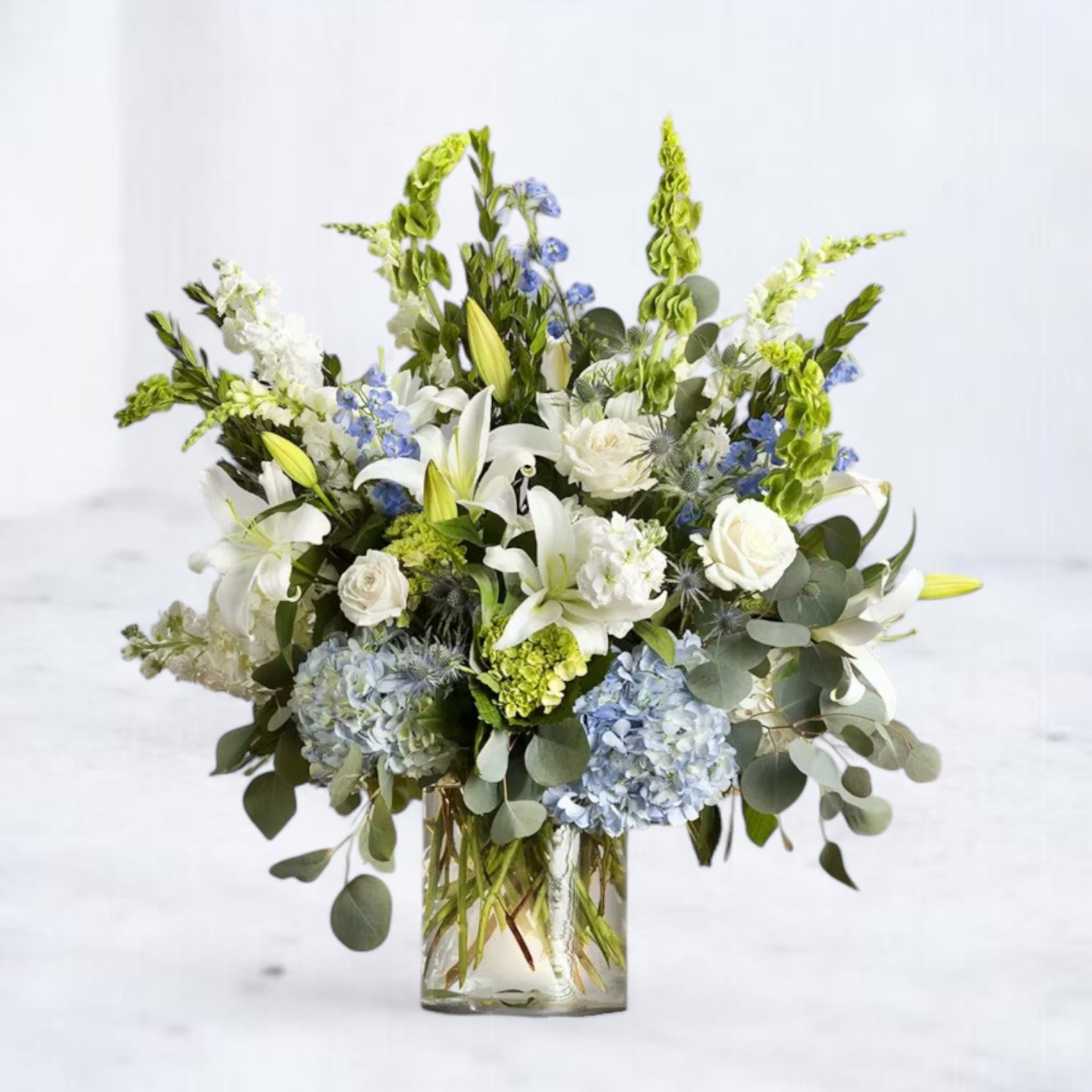 Tranquil Horizon Bouquet Tranquil Horizon Bouquet - Elevate your space with the serene beauty of the Tranquil Horizon Bouquet, a breathtaking arrangement inspired by the calm and refreshing hues of the sky and sea. Featuring soft blue hydrangeas, pristine white lilies, roses, and bells of Ireland, this lush design is complemented by delicate delphinium and textured greenery. Artfully arranged in a clear glass vase, this bouquet is perfect for expressing peace, comfort, and elegance for any occasion, from heartfelt condolences to joyful celebrations.